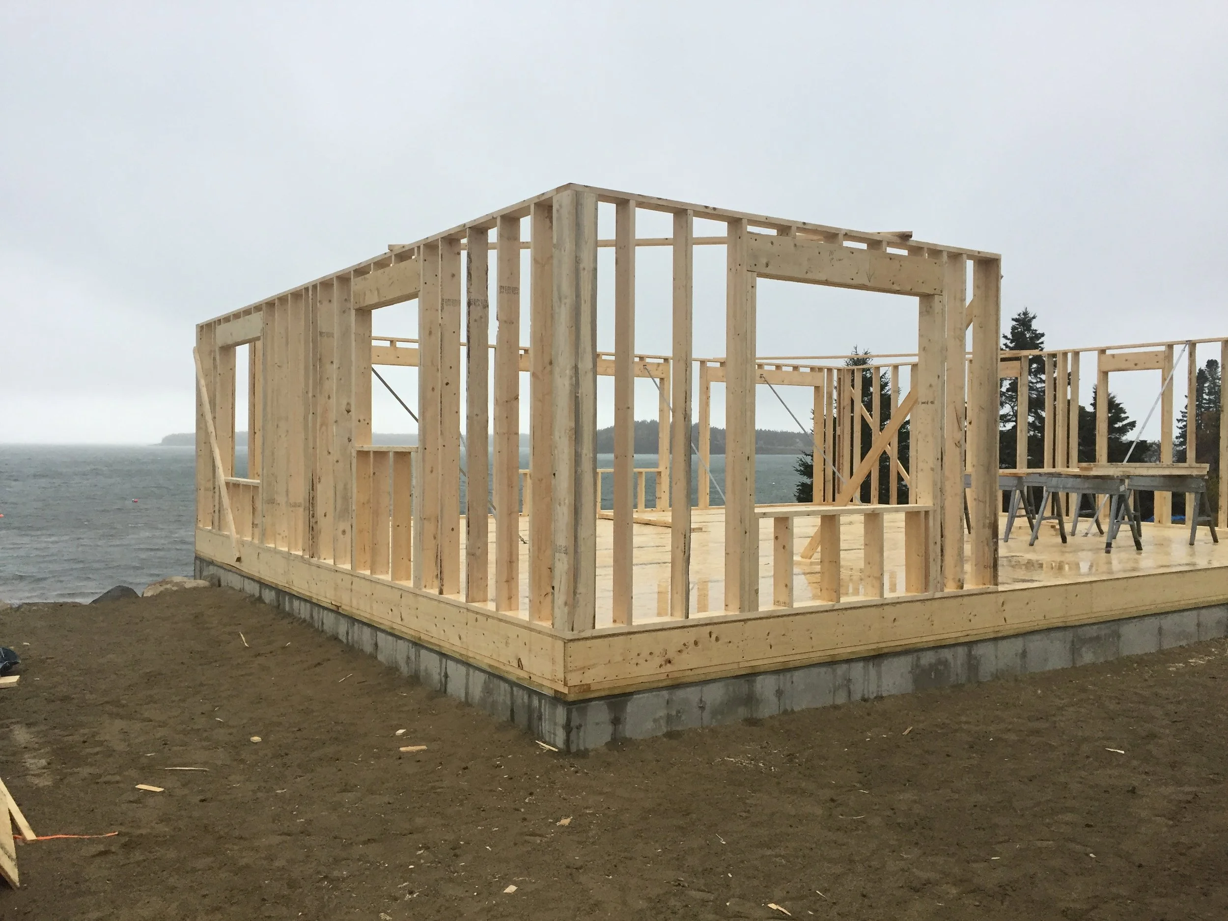 Wooden framing of a house under construction on a foundation near the water, with overcast sky.