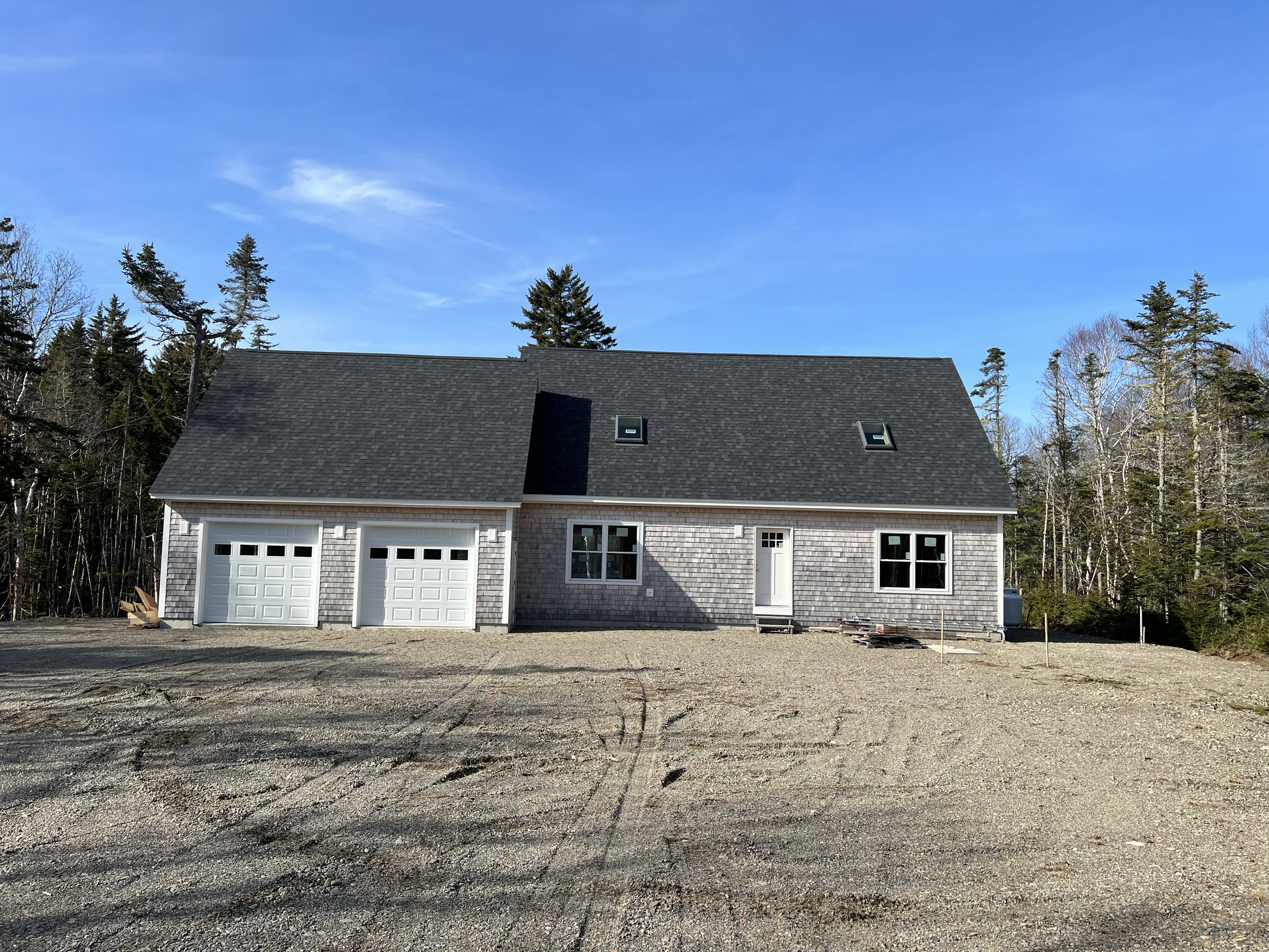 Newly constructed house with garage, gray shingle siding, black roof, windows, door, and surrounded by bare dirt and trees in the background.