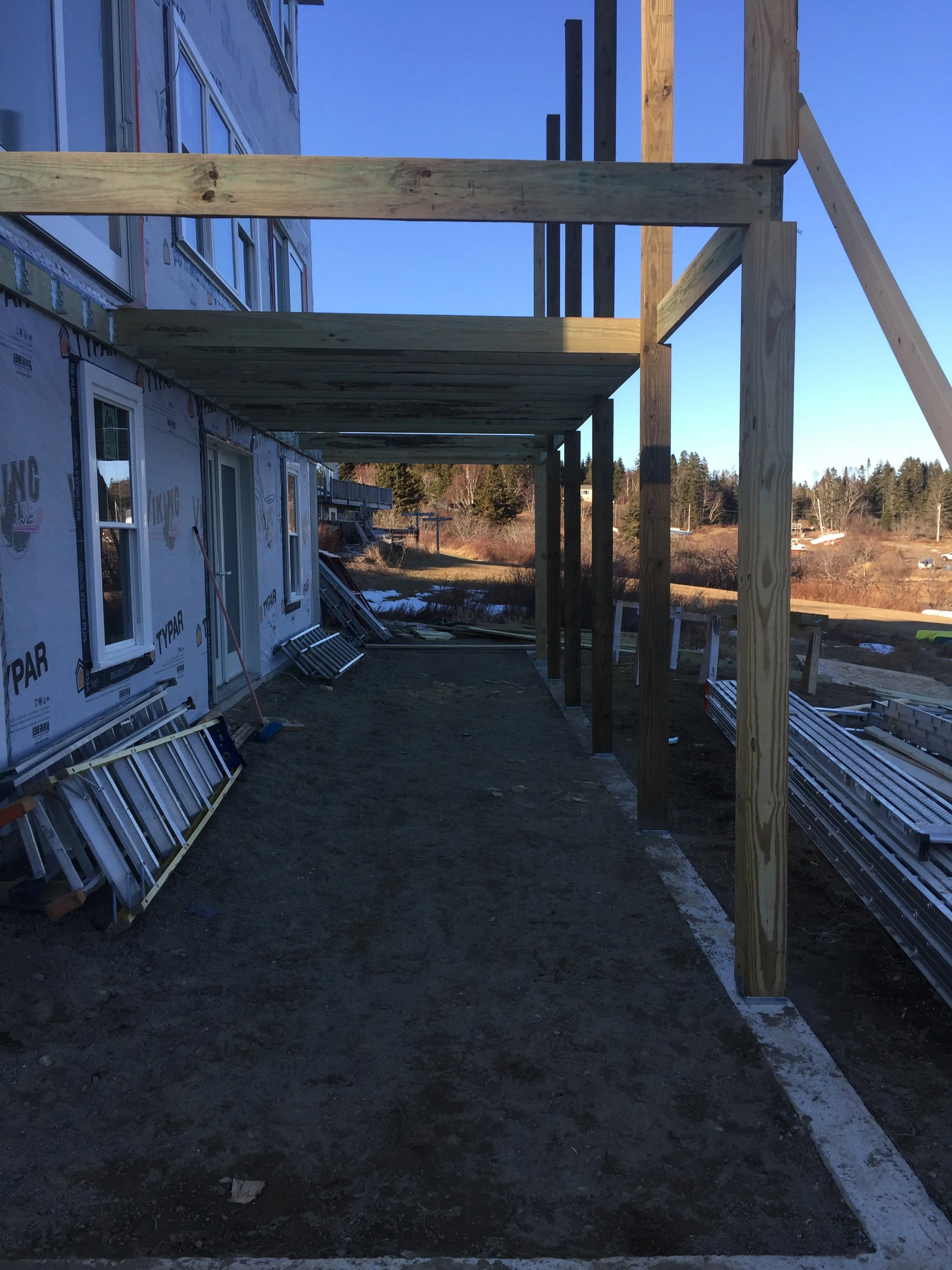 Construction site showing the framework of a building with wooden beams and a patio or balcony under construction. Piles of siding and construction materials are on the ground outside.