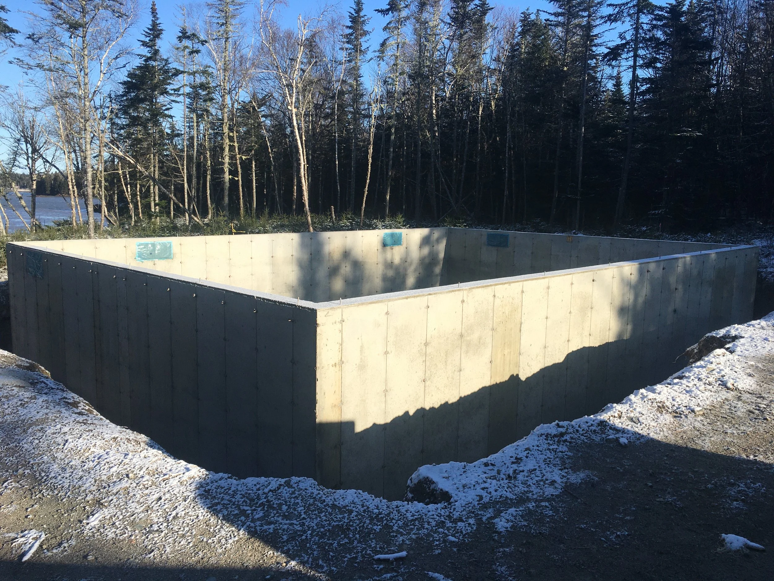 Concrete foundation in construction site surrounded by snow, with trees and water in the background.