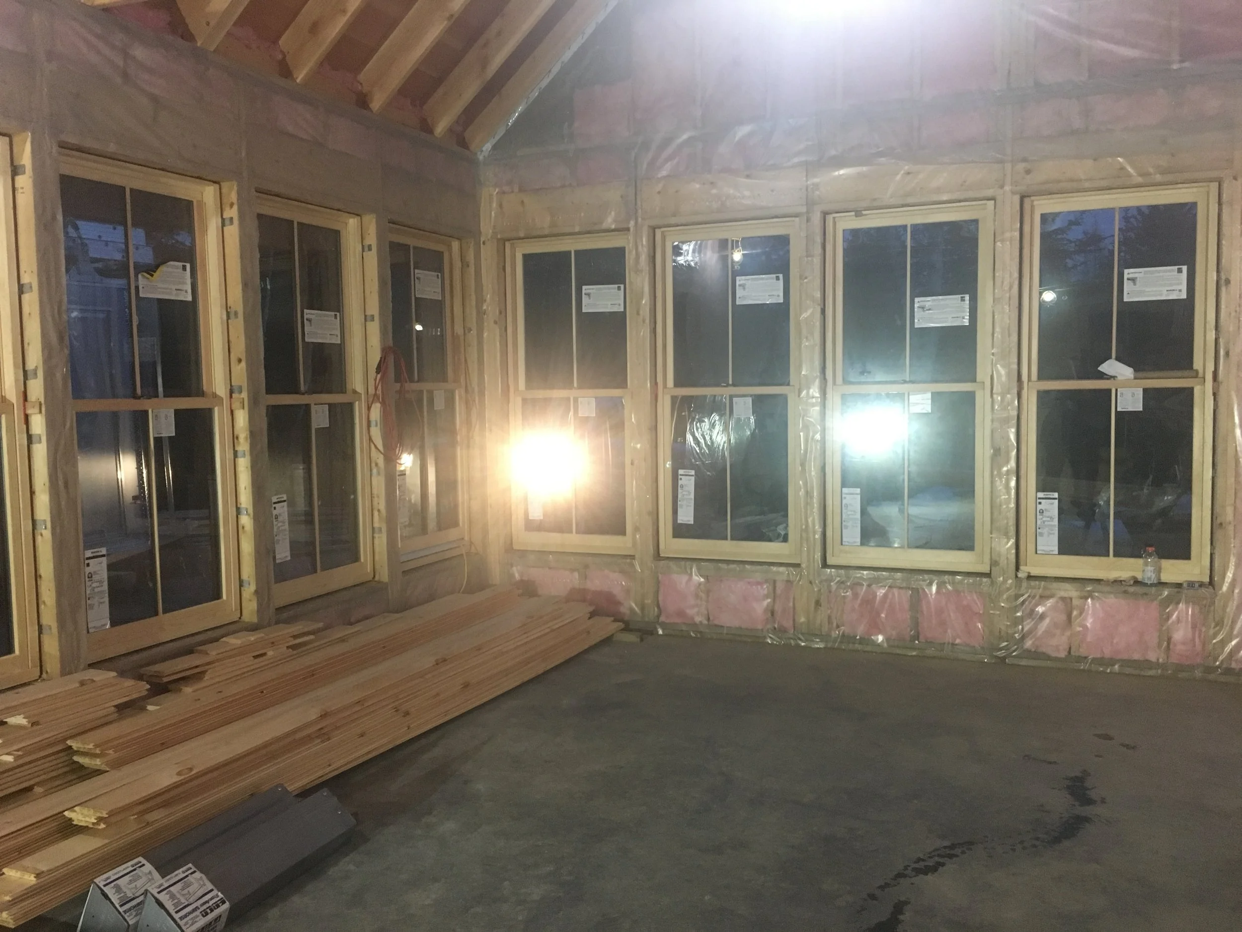 Interior of a house under construction with multiple window frames installed, some wood planks on the floor, and insulation material visible around the windows.