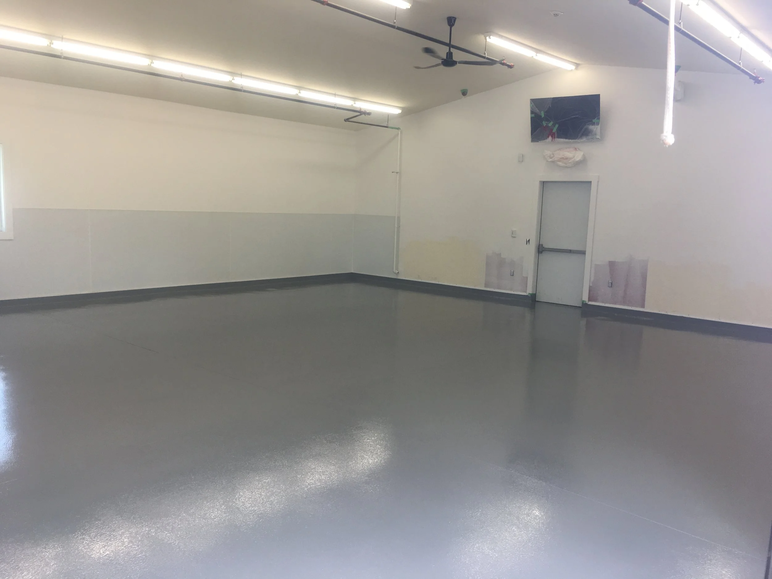 Empty industrial or commercial space with polished concrete floors, white walls, fluorescent lighting, and a mounted television or monitor on the wall.