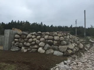 A pile of large rocks and stones along a rural landscape with trees and power lines in the background under a cloudy sky.