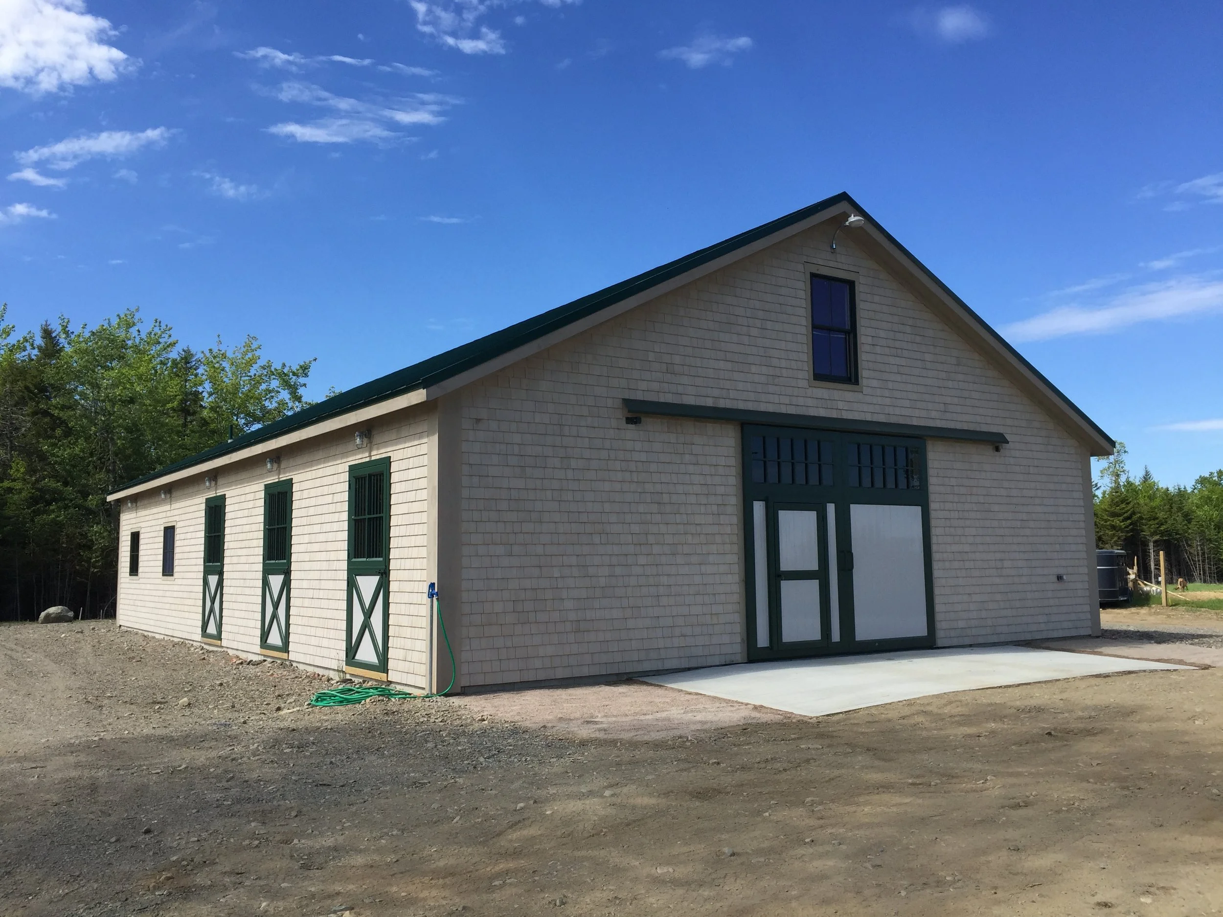 New beige barn with green doors and trim under a blue sky, surrounded by trees.