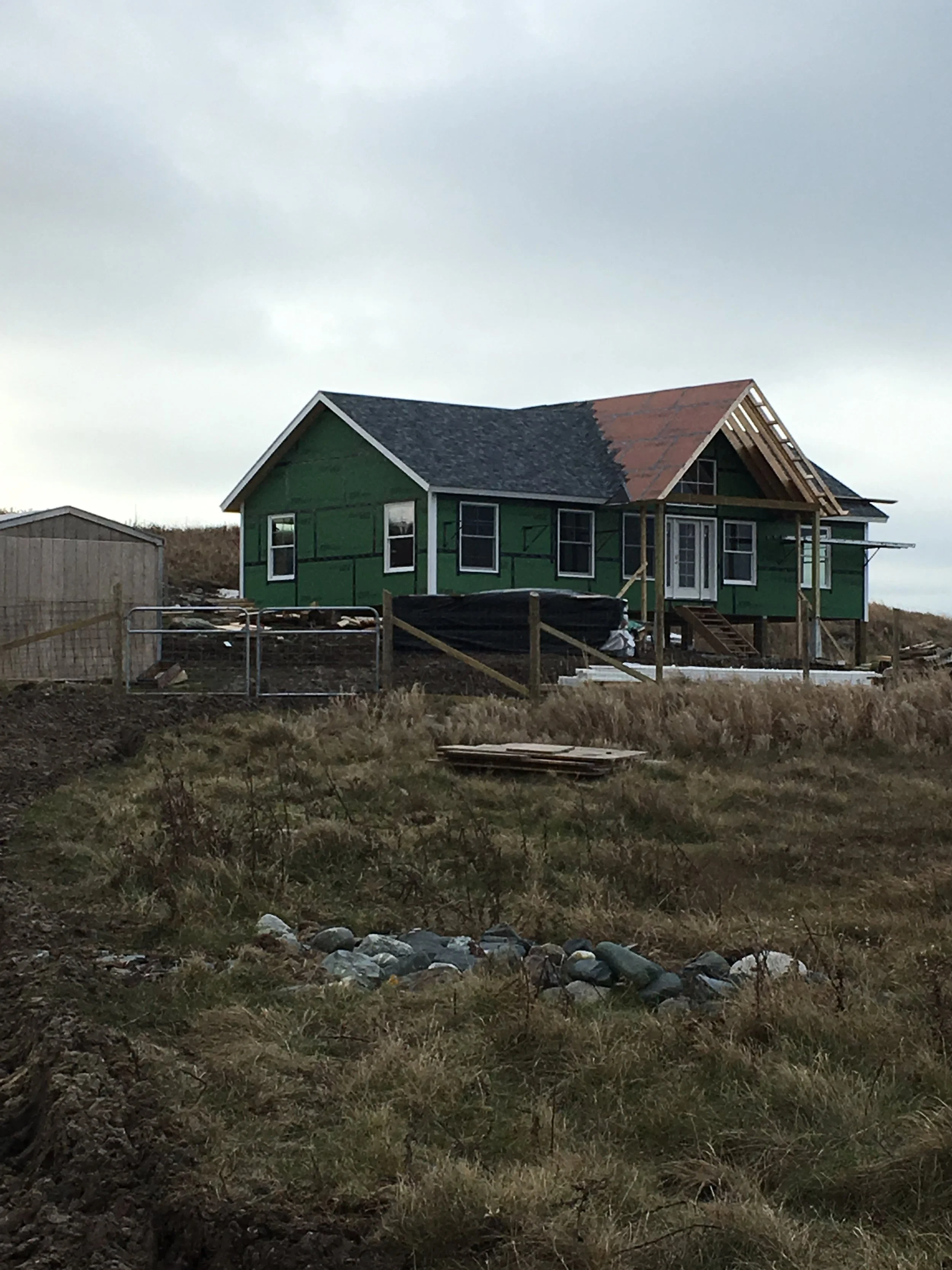 New house under construction with green exterior, partially built roof, and surrounding construction materials on a grassy plot.