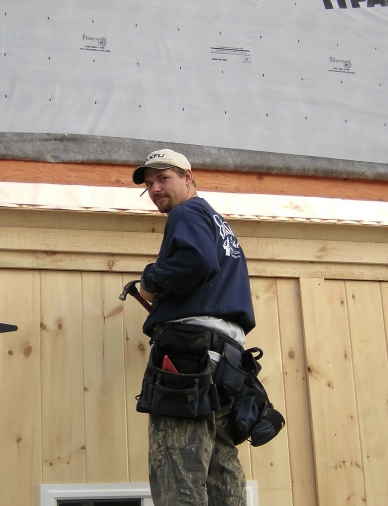 A construction worker standing indoors in front of a unfinished wooden wall with a large insulation sheet overhead. The worker is wearing camouflage pants, a dark work jacket, a tool belt, and a beige cap.