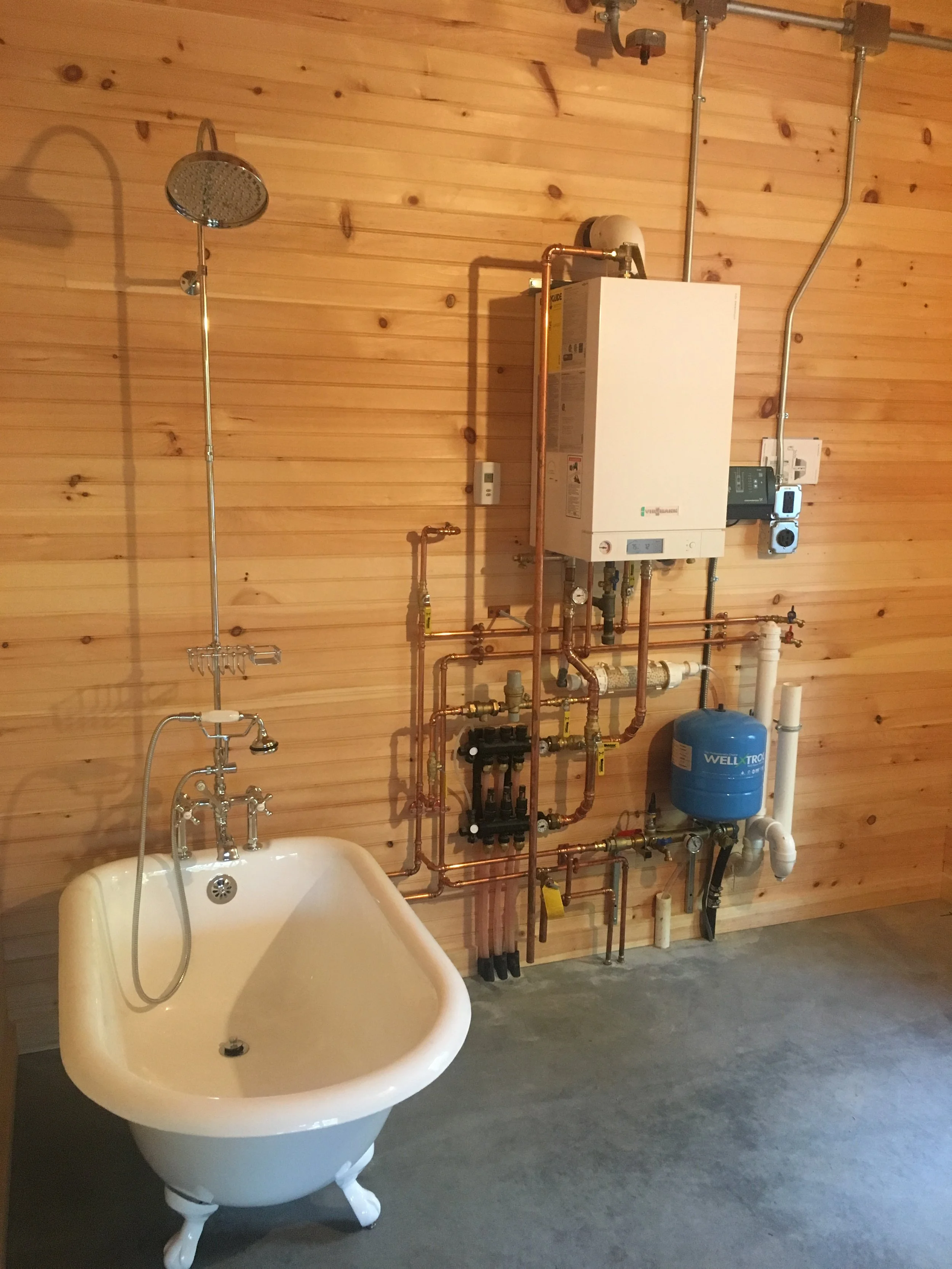 A bathroom space with a vintage clawfoot bathtub, a wooden wall with plumbing and heating equipment, including pipes, a water heater, and a pressure tank, mounted on the wall.