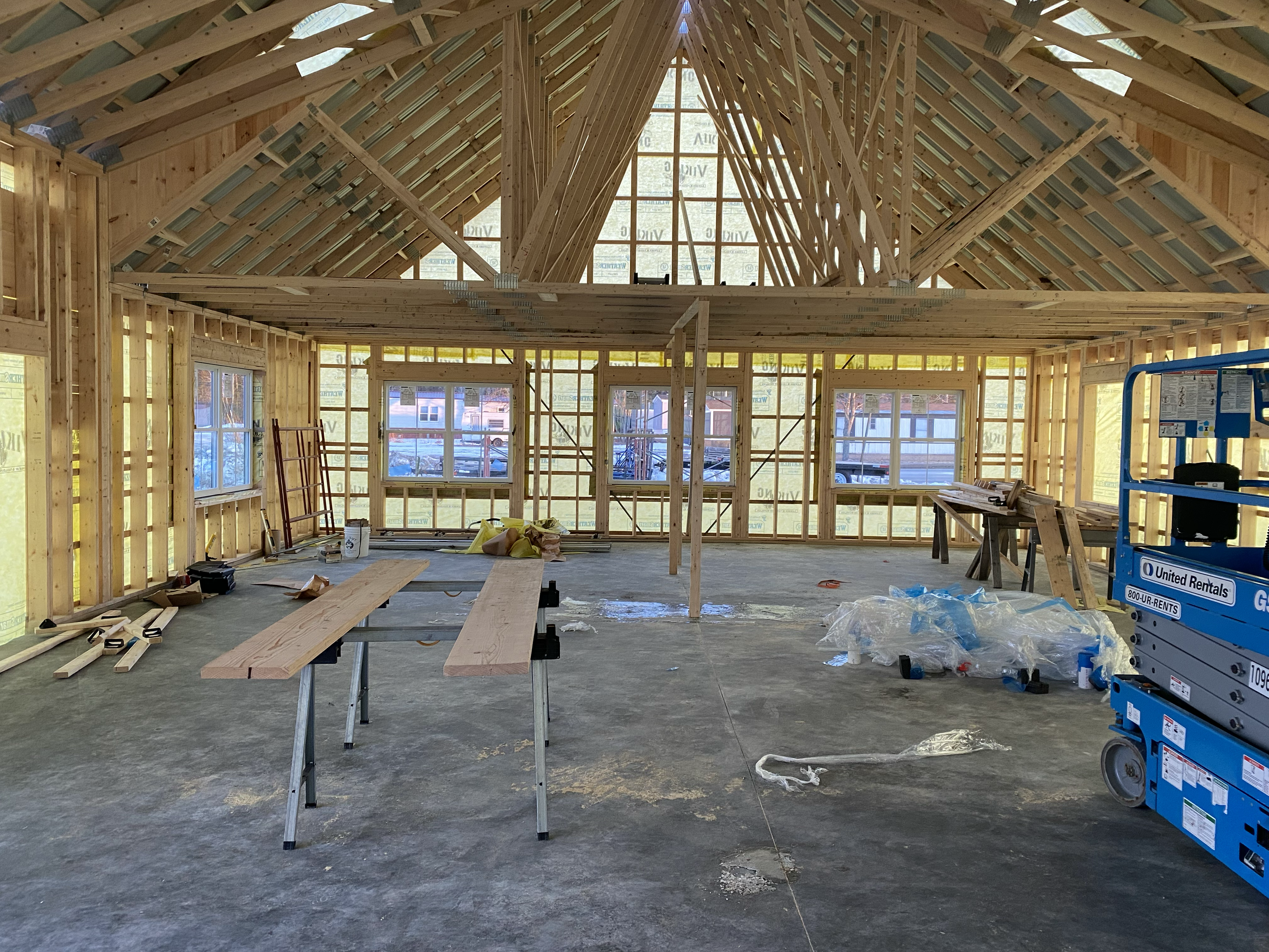 Interior view of a building under construction with wooden framing, large windows, and a high vaulted ceiling, with construction tools and materials scattered on the concrete floor.