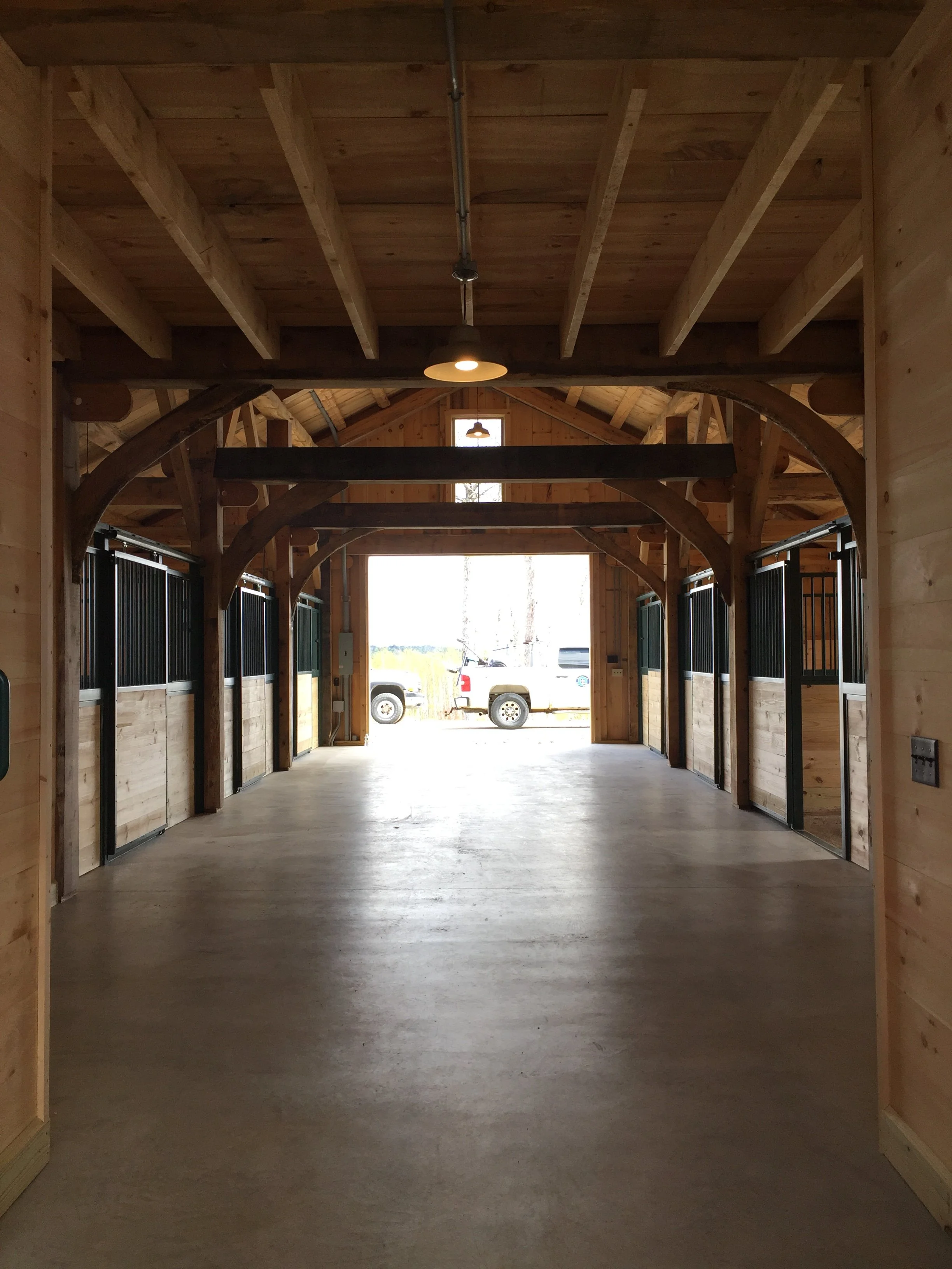 Interior view of a horse stable with wooden walls, stalls on either side, and a large open doorway at the end showing trucks and trees outside.