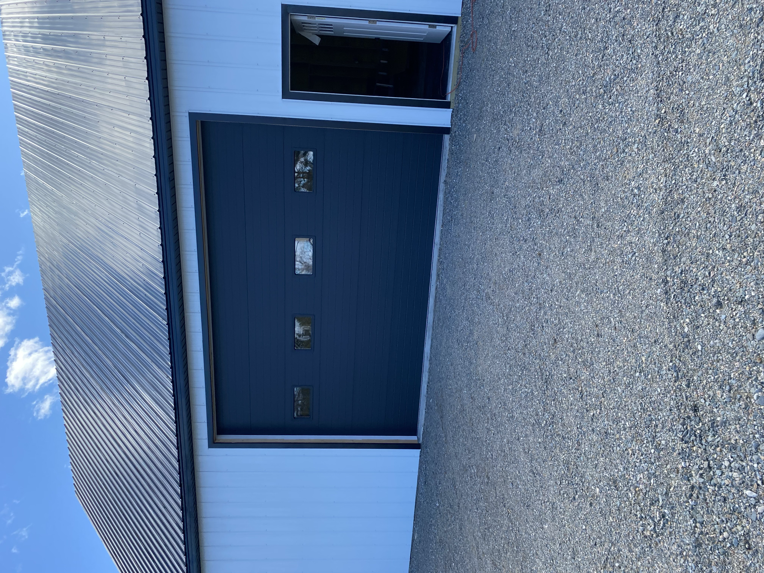 A partly open garage door on a white building with a gravel driveway in front, under a blue sky.