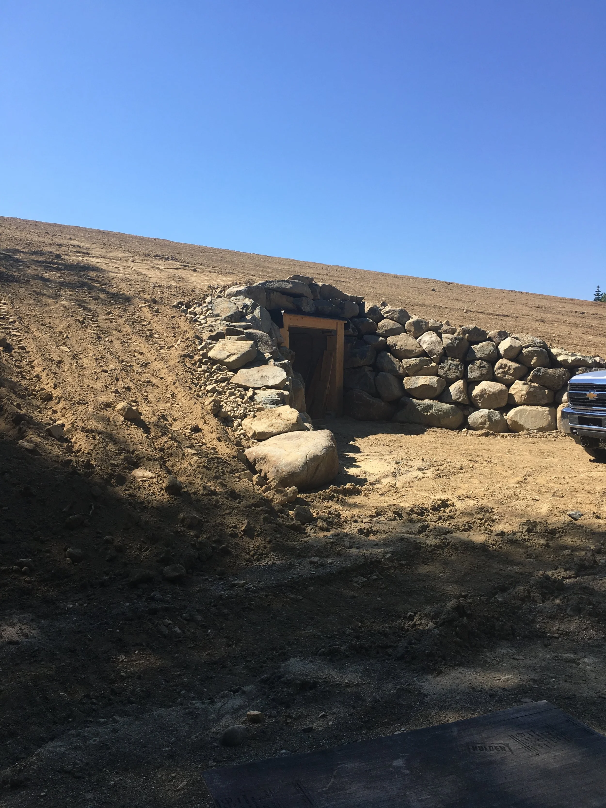 A dirt road leads to a tunnel entrance built into a hillside with large rocks surrounding it. A vehicle is partially visible on the right side of the image.
