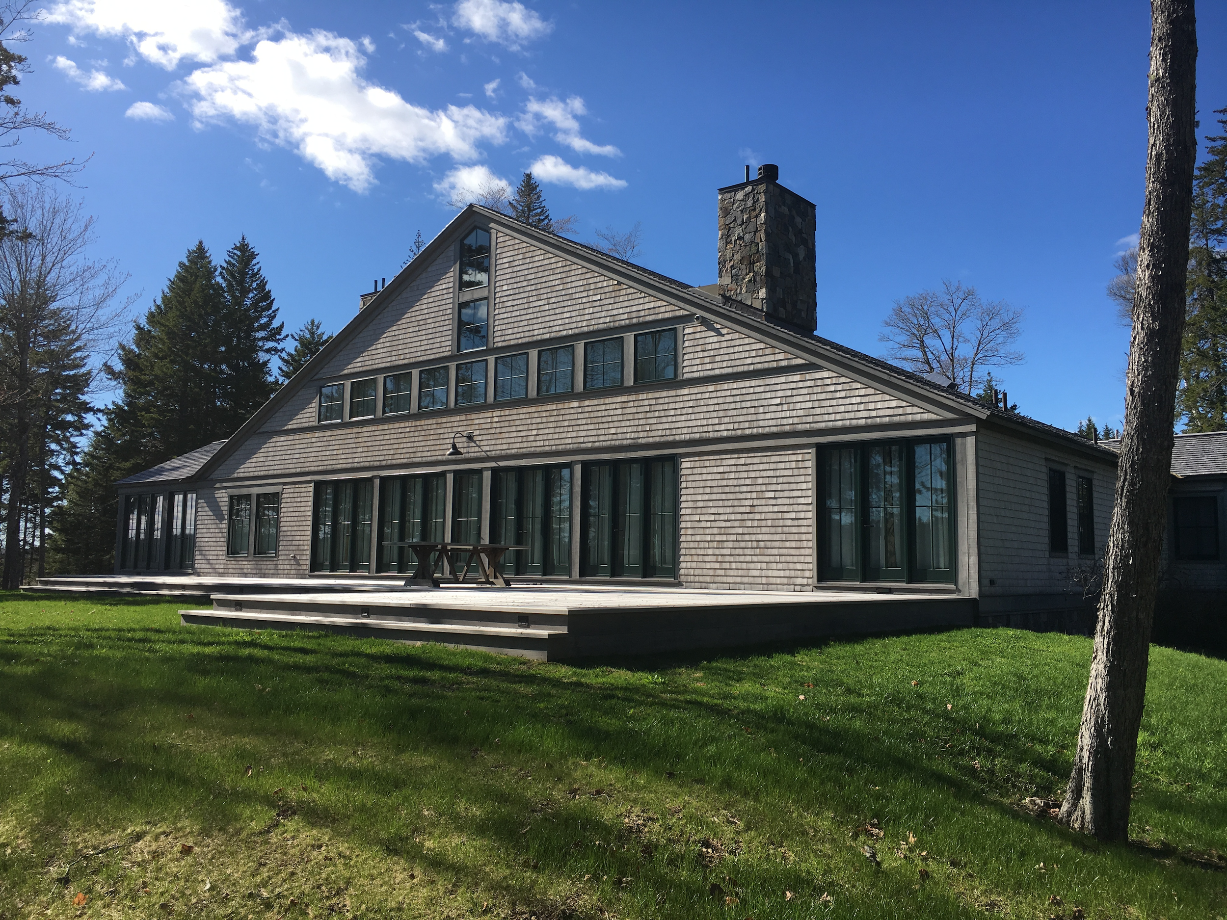 A large house with a gray exterior, multiple glass doors, and a wooden deck in front, surrounded by grass and tall trees under a partly cloudy blue sky.