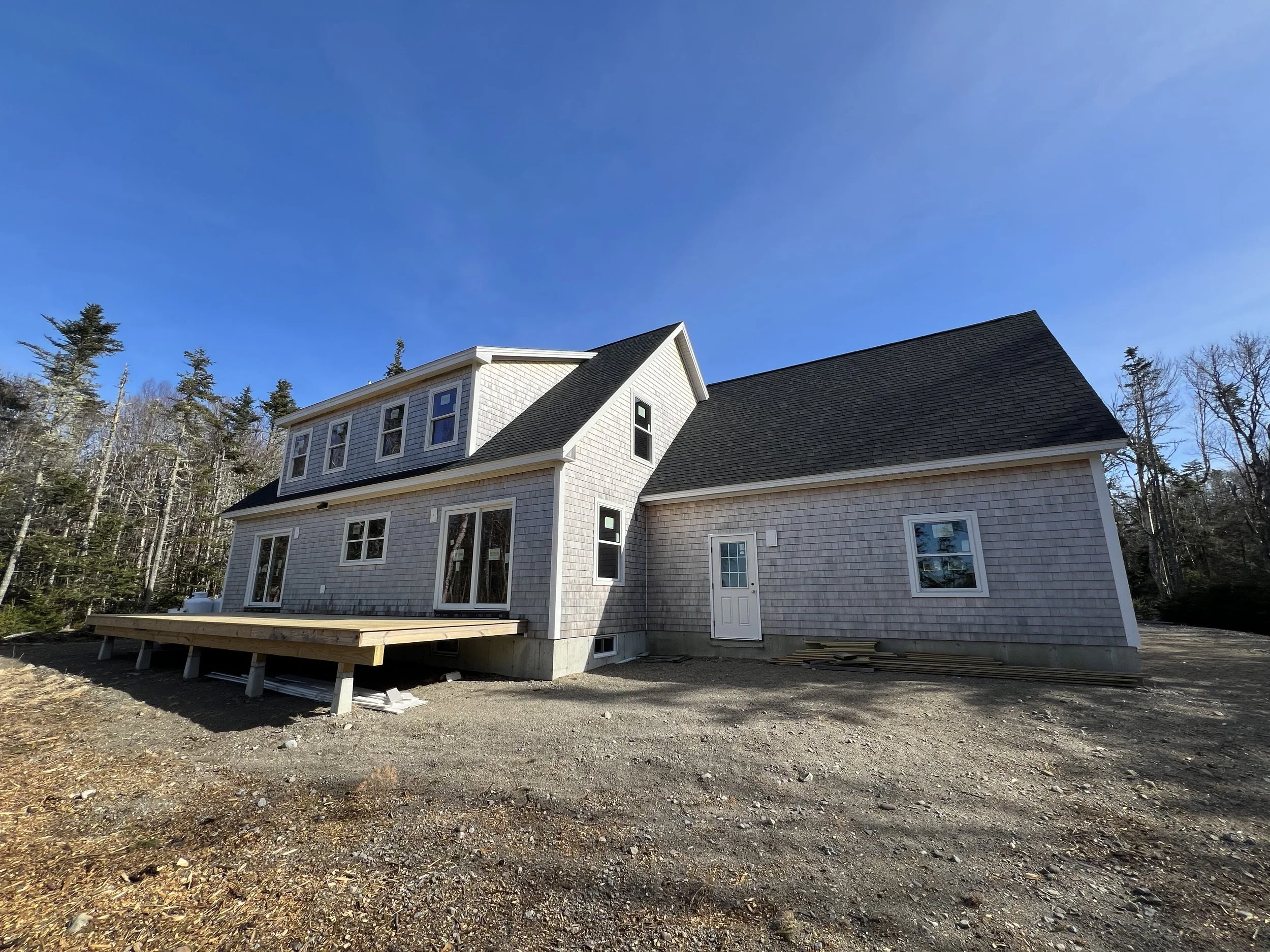 A new two-story house under construction with a wooden deck, surrounded by a dirt yard and trees in the background, under a clear blue sky.
