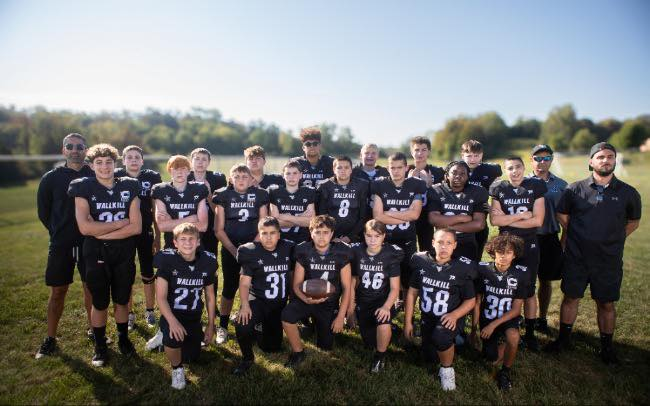 A youth football team posing outdoors on a grassy field with coaches, wearing black jerseys with white text and numbers.