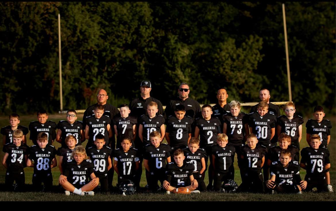 Youth football team in black uniforms, team photo outdoors with coaches behind, football field, trees in background.