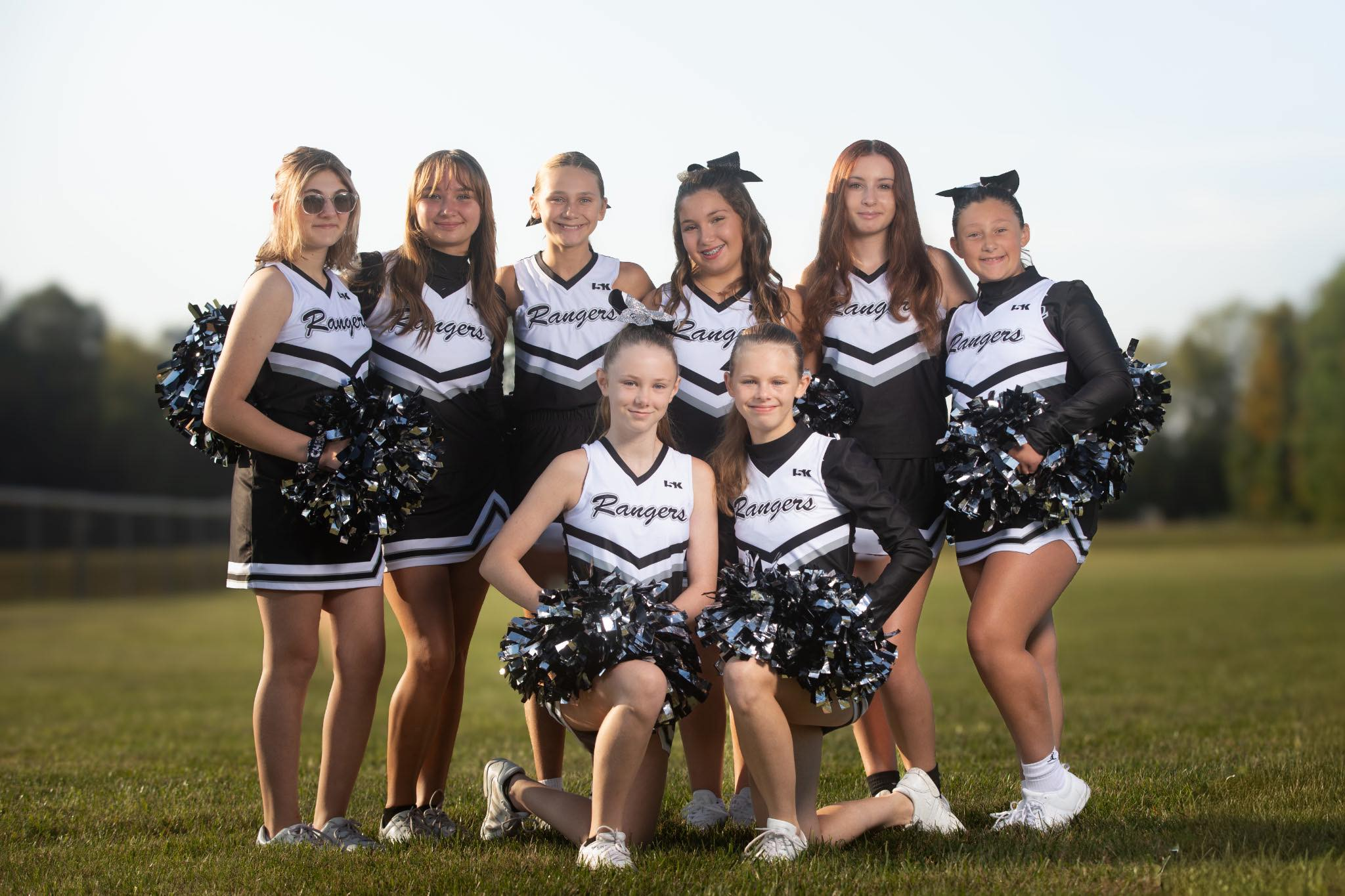 A group of eight young cheerleaders dressed in black and white uniforms, holding black and silver pom-poms, posing outdoors on a grassy field with a blurred background and a clear sky.
