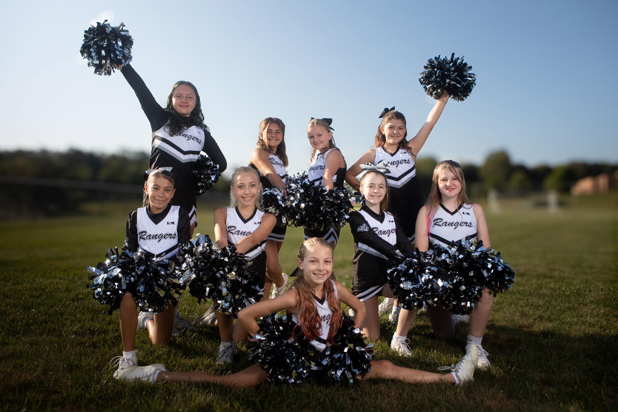 Group of young cheerleaders in black and white uniforms posing outdoors on grass field, some holding pom-poms, with a clear blue sky in the background.