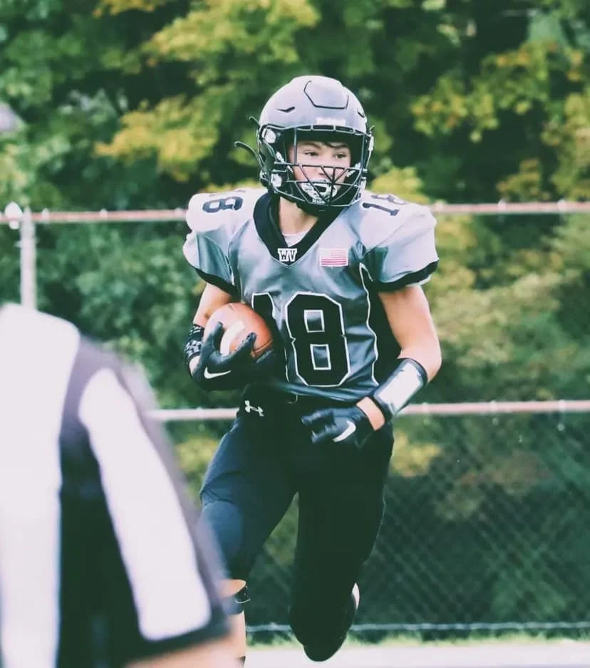 Young football player in a gray uniform holding a football, running on the field with a helmet and gloves, during a game.