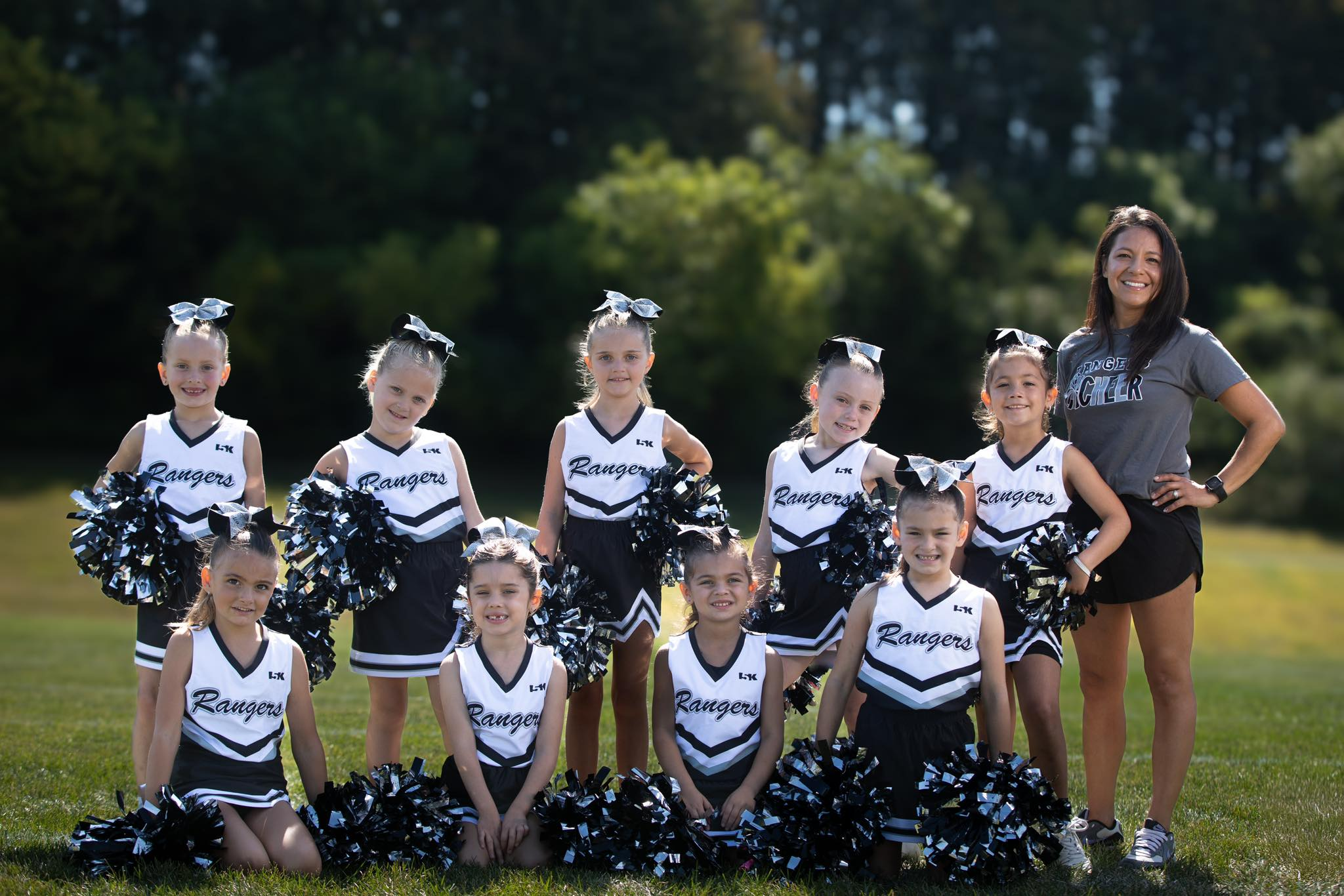 A group of young female cheerleaders with black and white uniforms labeled 'Rangers', holding black and white pom-poms, standing outdoors on a grass field with trees in the background, accompanied by an adult woman coach wearing a gray shirt that say