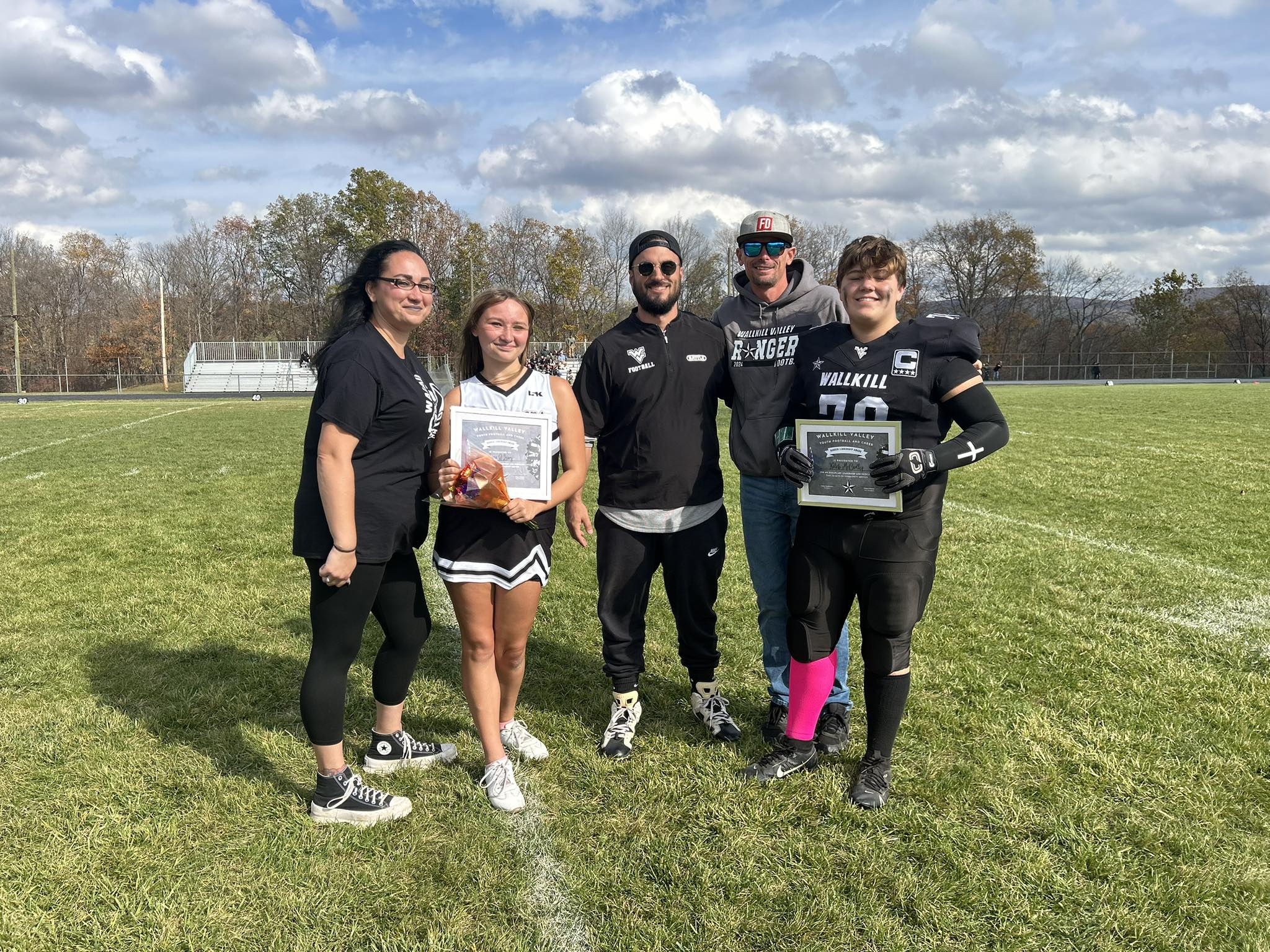 Group of five people standing on a sports field, holding awards, with trees and a cloudy sky in the background.