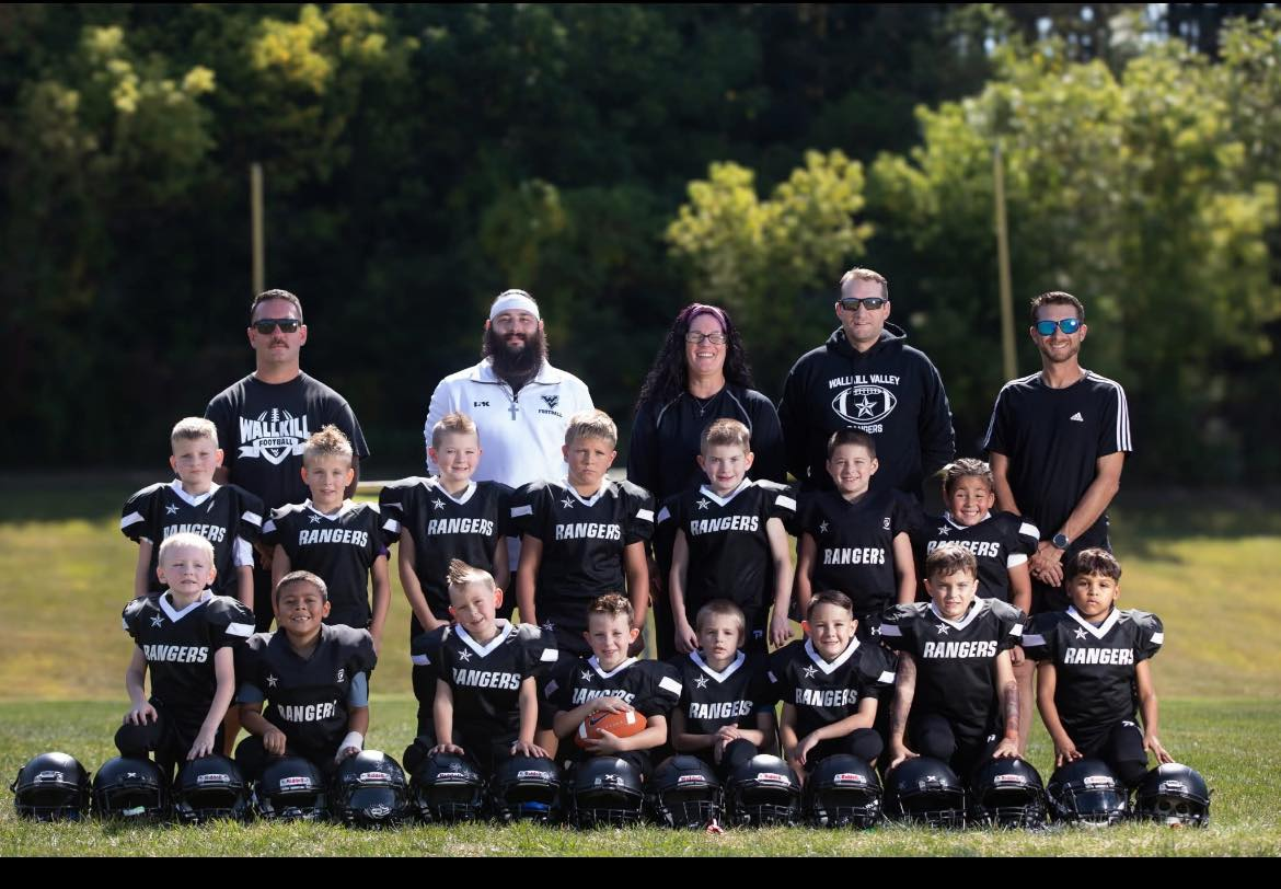 A youth football team in black jerseys with 'RANGERS' written on them, along with their coaches and staff, posing outdoors on a grassy field with trees in the background. Helmets are lined up in front of the kids, and one kid holds a football.
