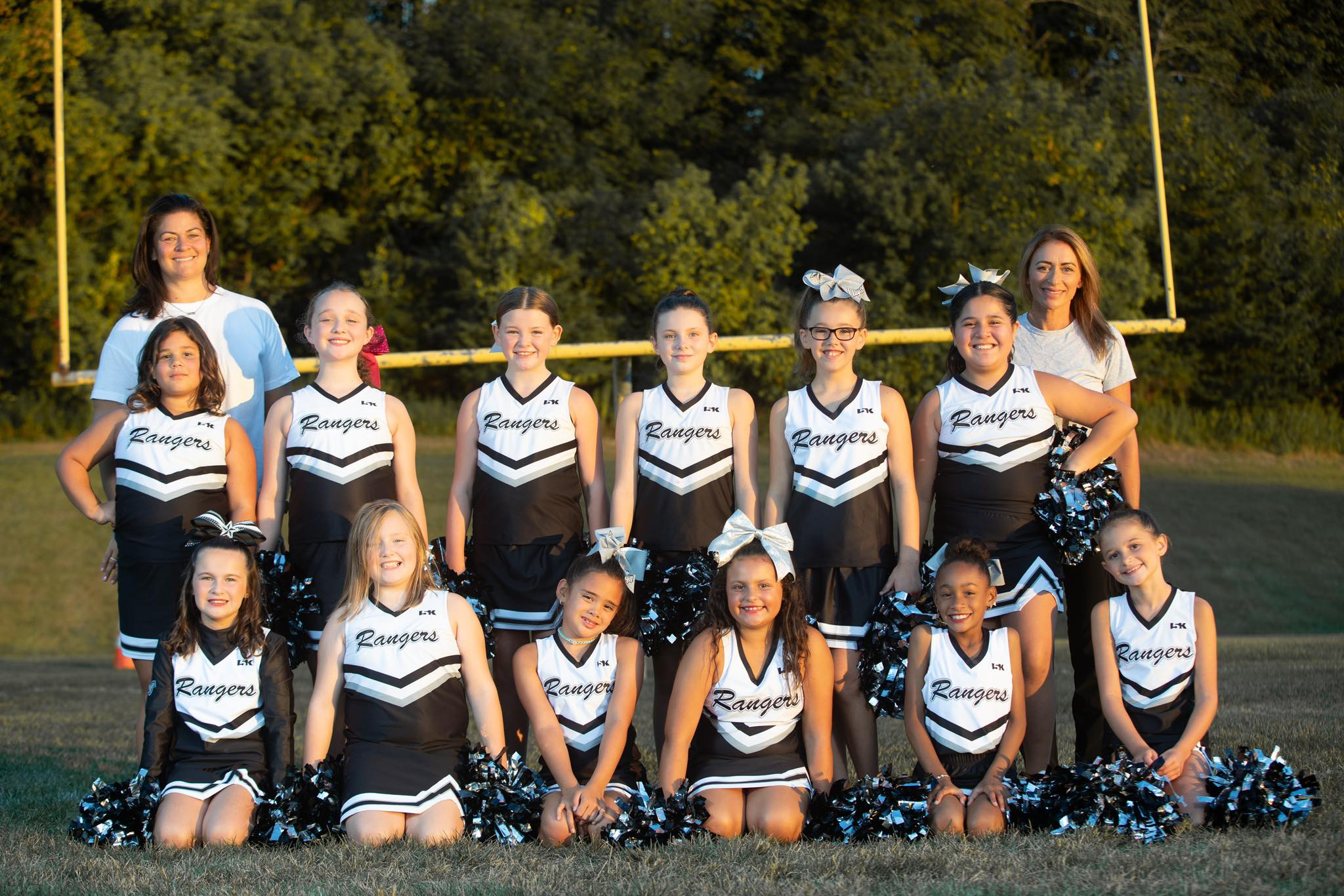 A group of young cheerleaders in black and white uniforms with 'Rangers' written on them, posing for a photo on a football field during sunset, with two adult women, possibly coaches, standing behind them.