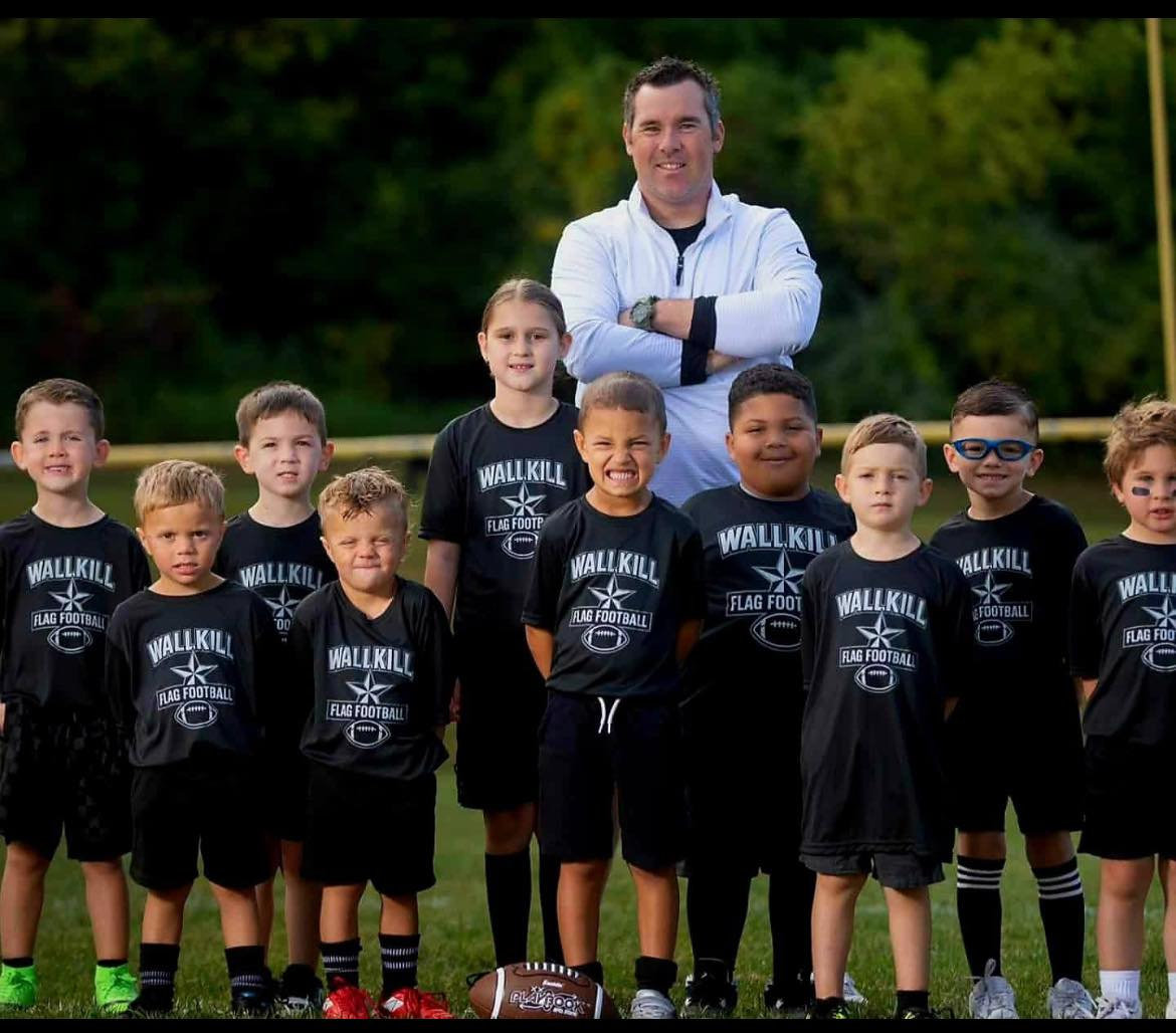 A youth flag football team with nine boys in black uniforms and their coach, standing on a football field outdoors with trees in the background. The coach is smiling with his arms crossed. Some boys are making faces, and a football is on the ground i