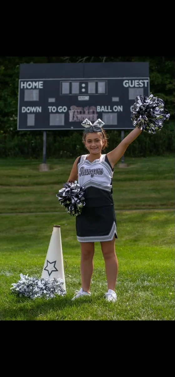 Young cheerleader in a black and white cheerleading uniform holding silver pom-poms, standing on grass with a scoreboard in the background, and a white cone with a star nearby.
