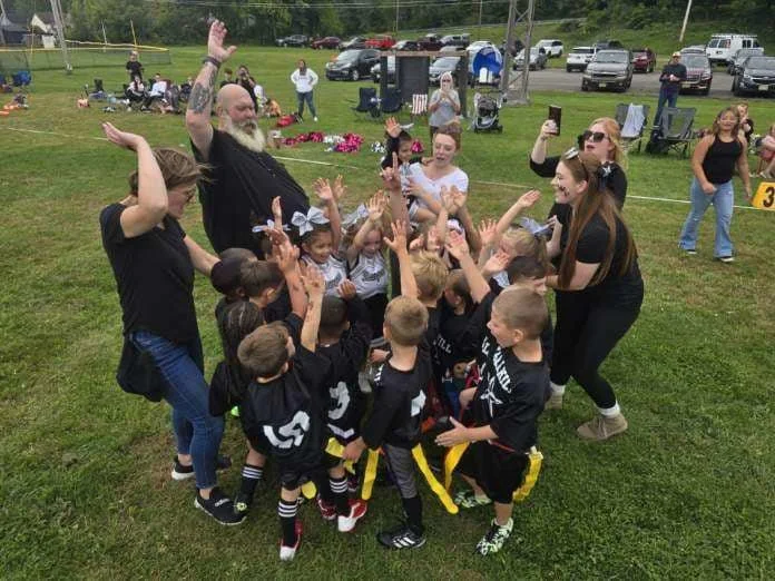 Group of children and adults celebrating outdoors, some children wearing football jerseys and yellow belts, with their hands raised, on a grassy field with parked cars and people in the background.