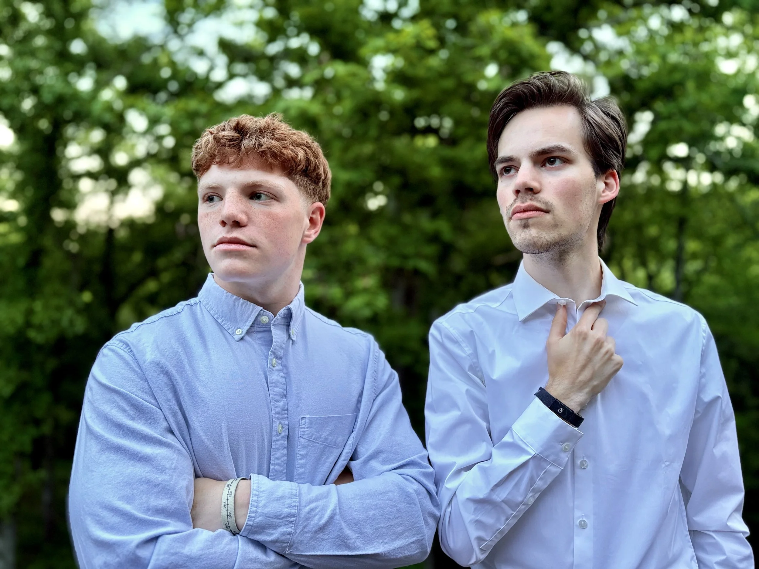 Two young men standing outdoors on a green, leafy background, dressed in light blue shirts, one with folded arms and the other adjusting his collar, both looking off into the distance.