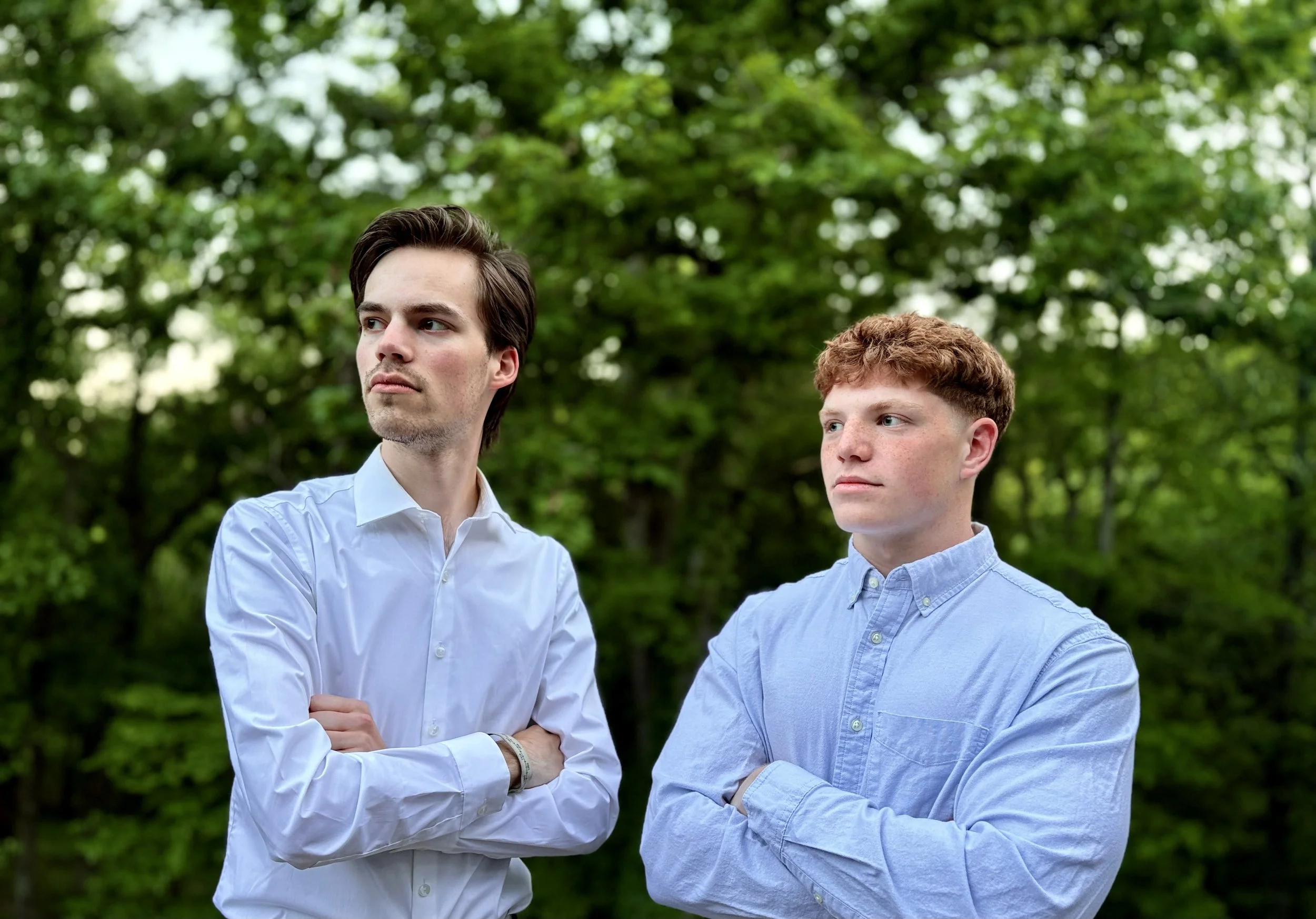Two young men standing outdoors in front of green trees, both wearing light blue shirts, one with crossed arms on the left and the other with arms at his sides on the right, looking in different directions.