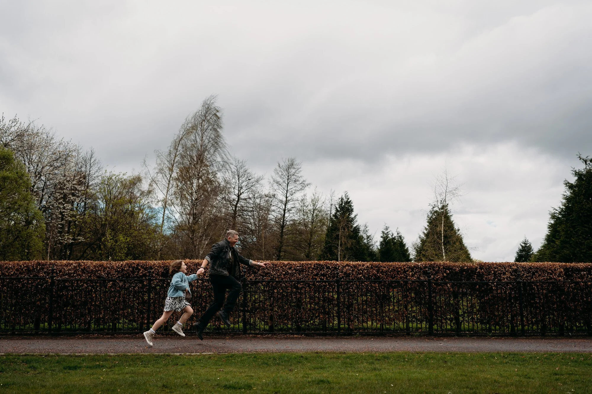 A man and a young girl are running hand in hand along a sidewalk bordered by a hedge. The man is smiling and wearing a dark jacket, and the girl is wearing a gray sweater and a patterned skirt. The background features a cloudy sky and trees without leaves.