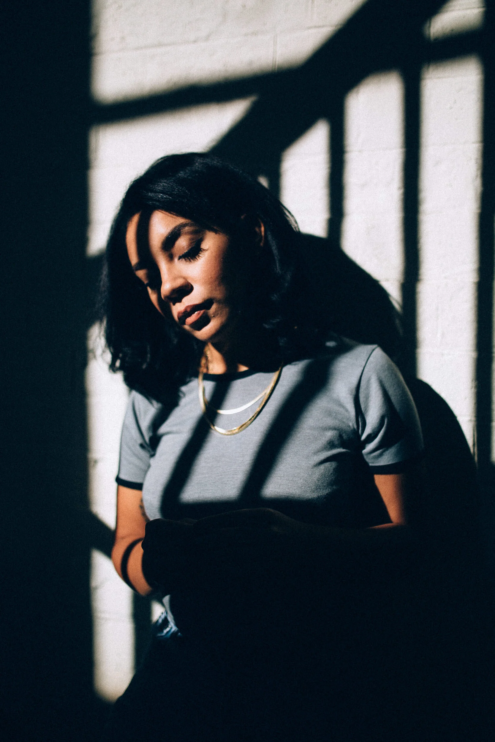A woman with dark hair and makeup, wearing a gray T-shirt, gold necklaces, and a black bracelet, stands against a shadowed backdrop of a white brick wall and cast shadows.