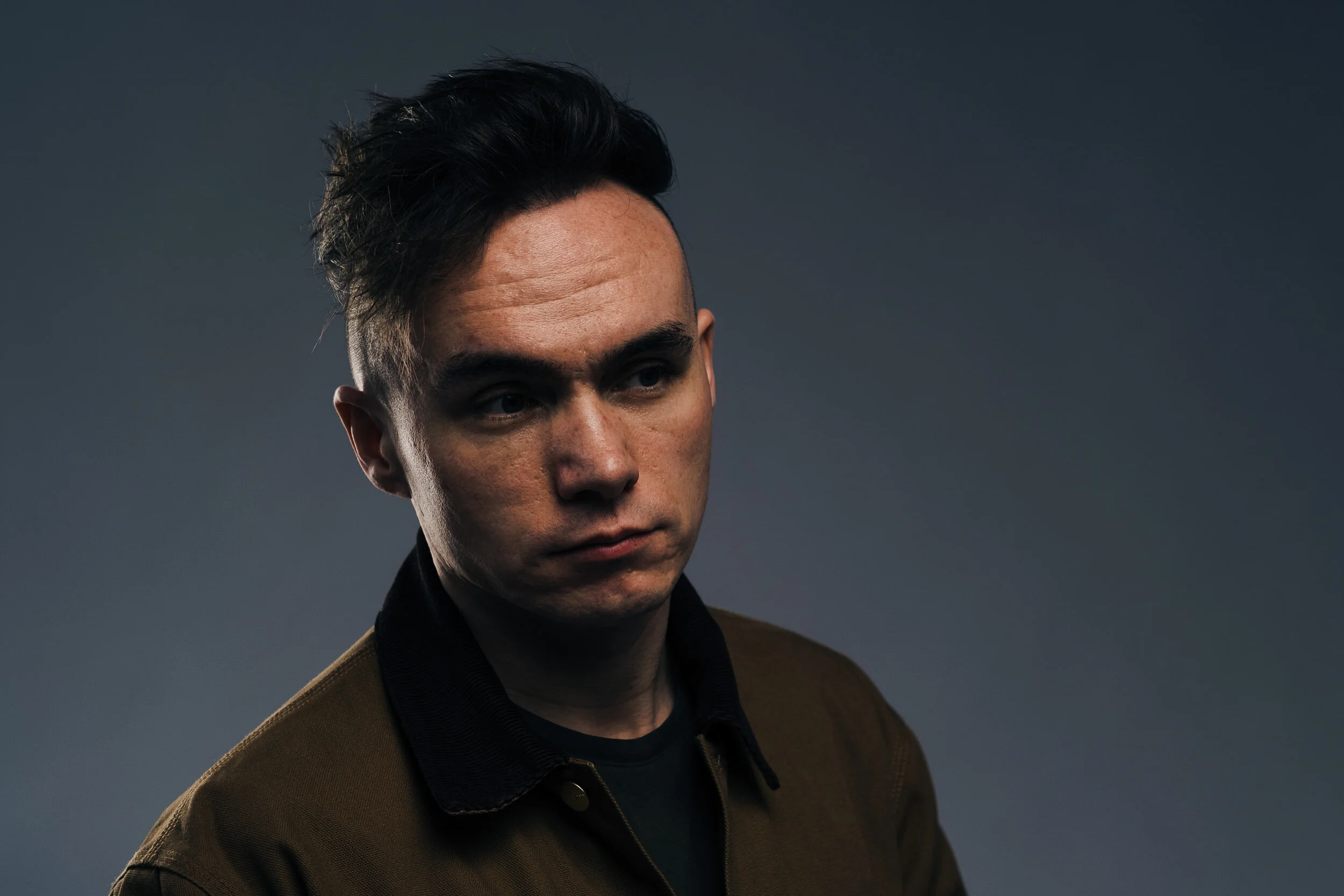 A man with dark hair styled upward, wearing a brown jacket over a black shirt, looking down and to the side against a plain gray background.