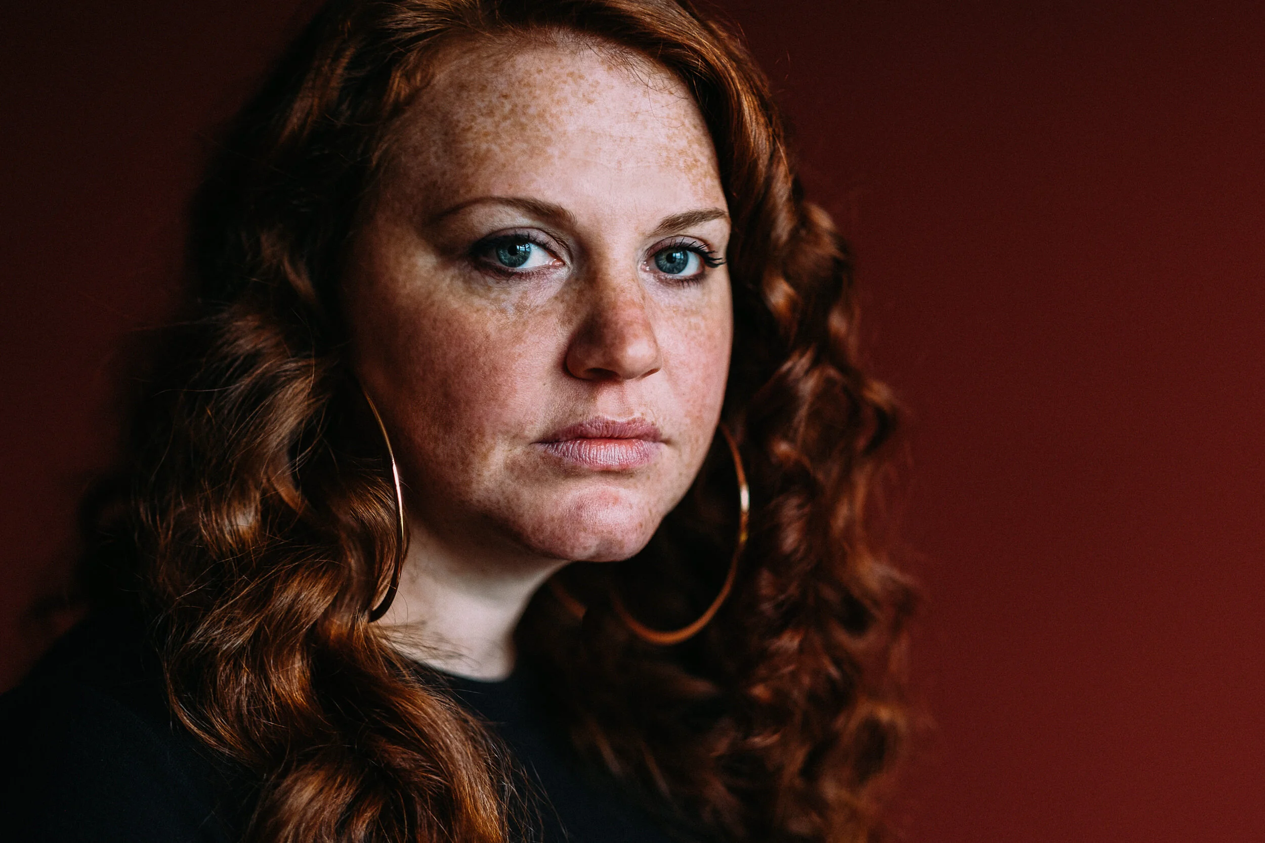 Close-up of a woman with red hair and blue eyes looking at the camera, with a dark background.