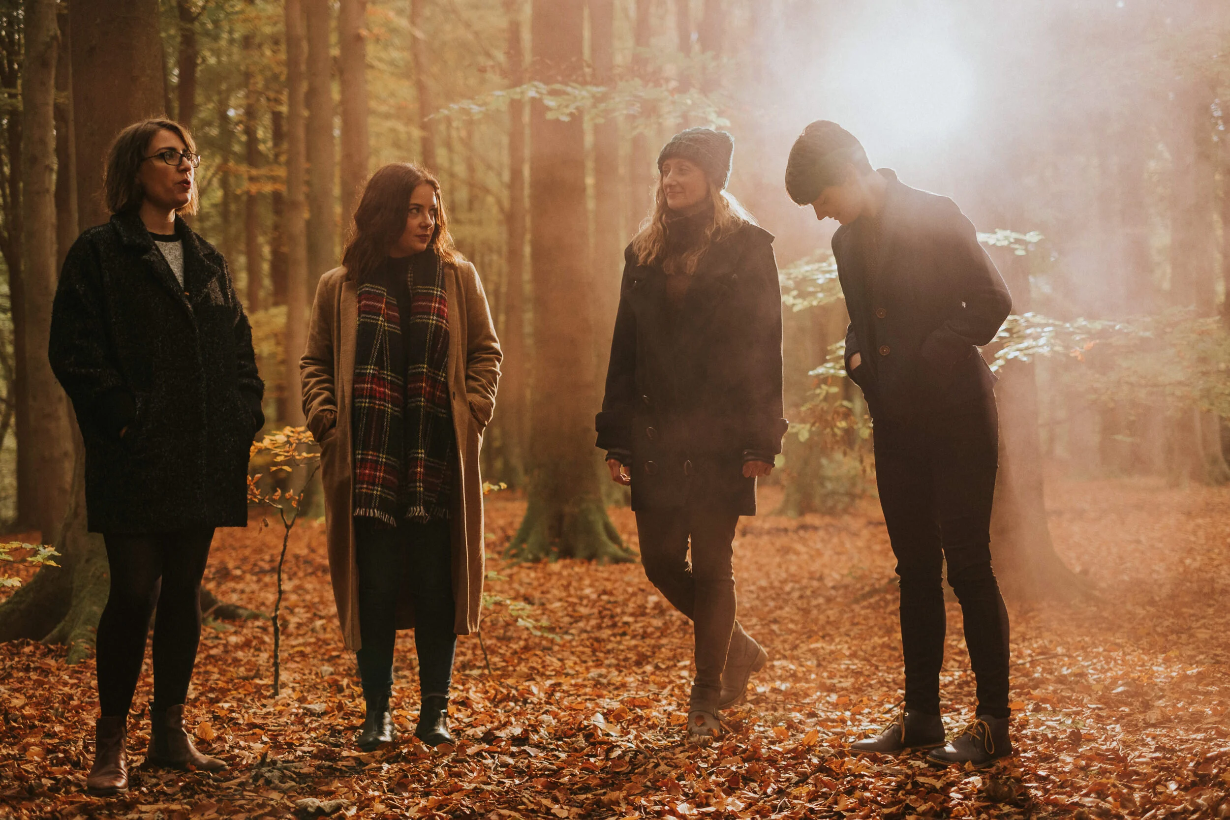 Four women standing and talking in an autumn forest with orange leaves on the ground, dressed warmly in jackets, scarves, and hats, illuminated by sunlight.