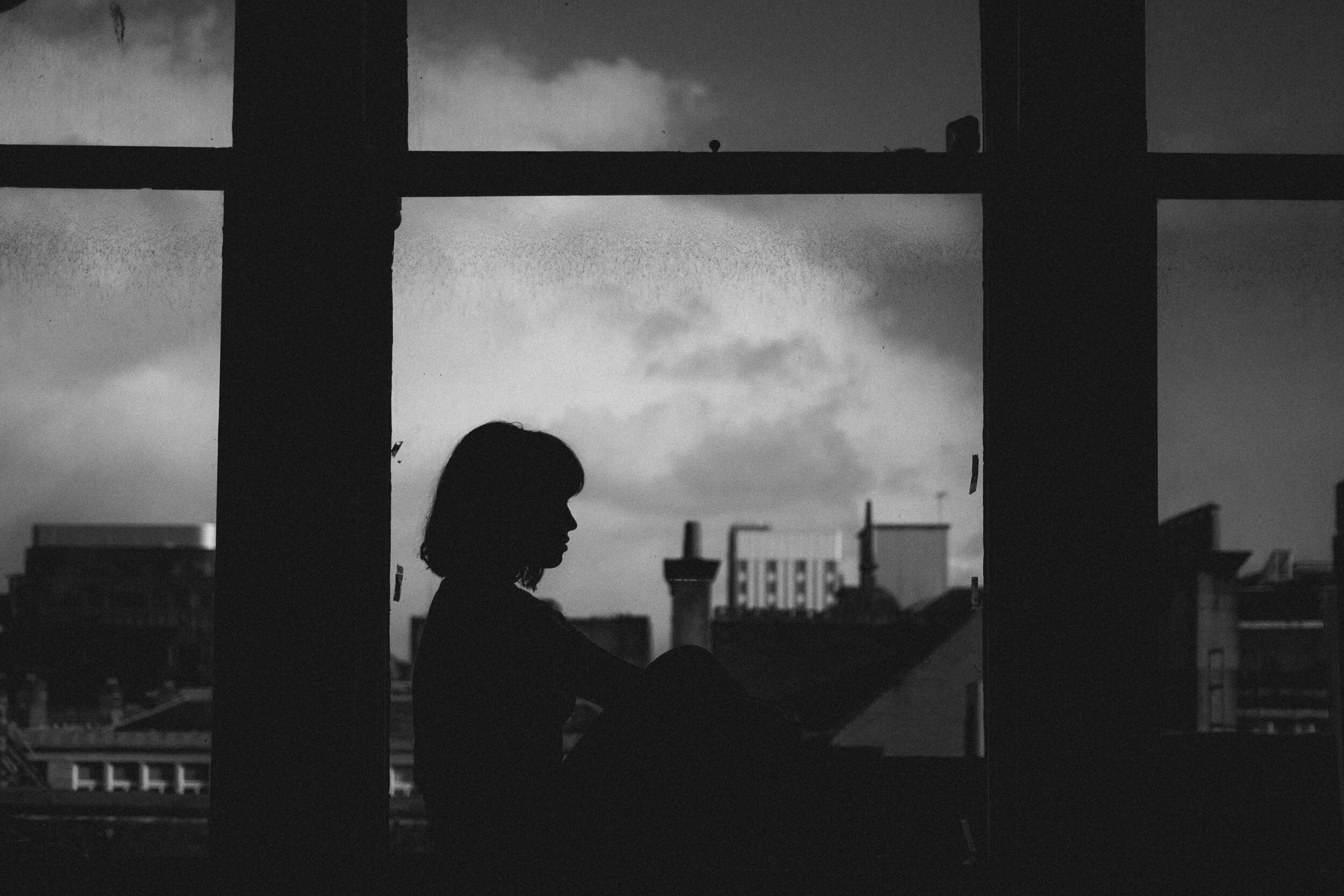 Silhouette of a person with curly hair sitting at a window, with an urban cityscape in the background during cloudy weather.