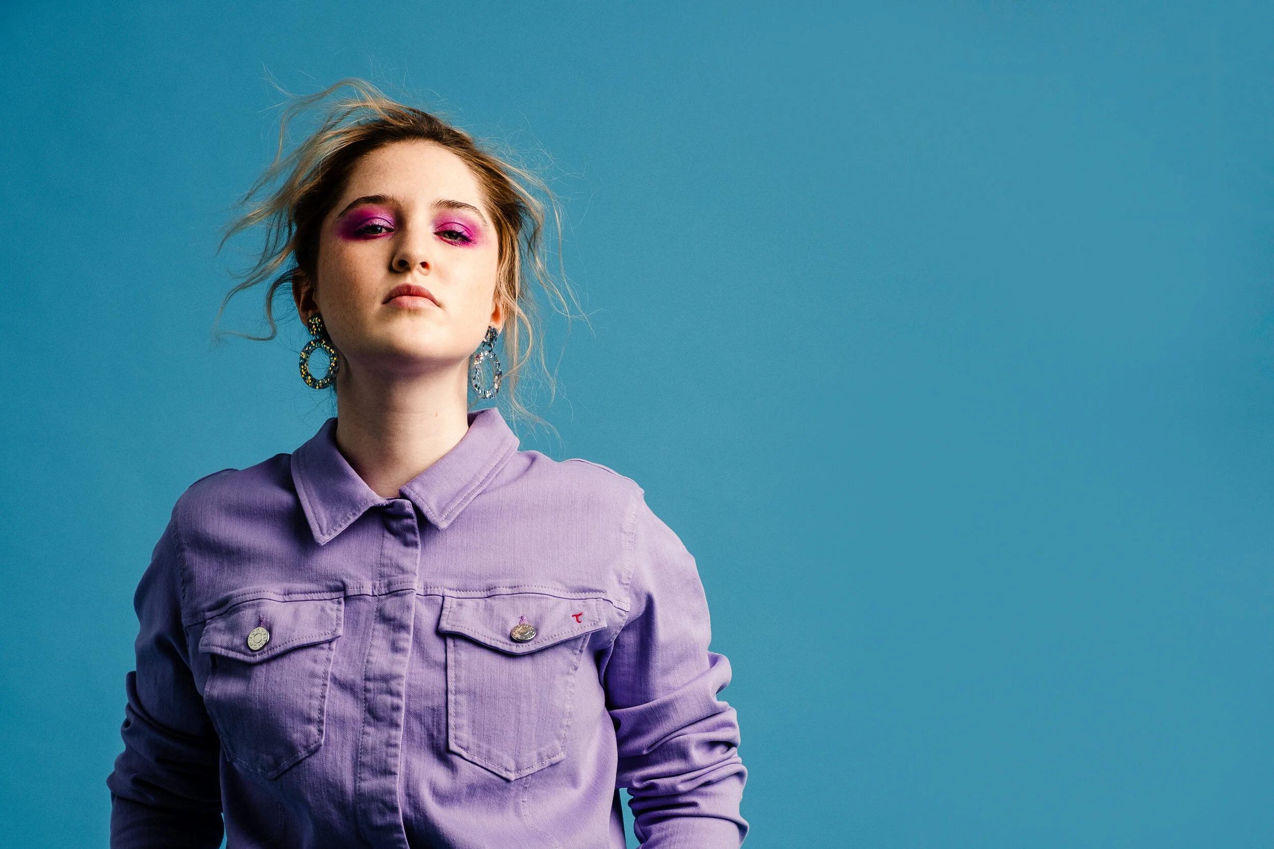 Portrait of a young woman with blonde hair, pink eyeshadow, large earrings, wearing a purple jacket, against a solid blue background.
