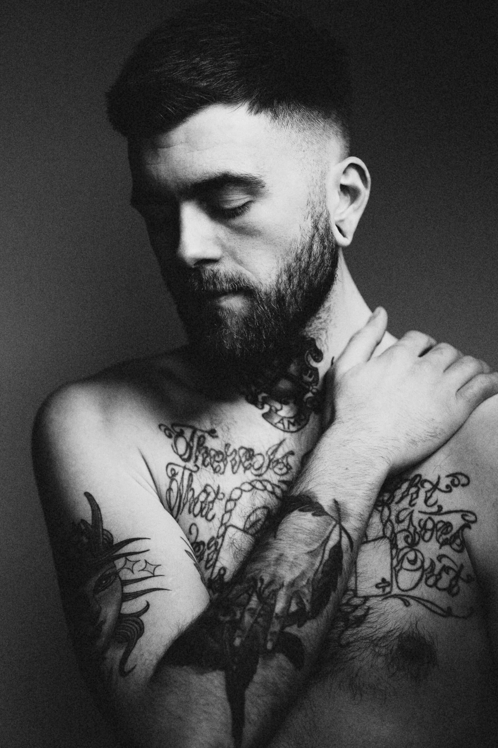 A black and white photograph of a shirtless man with tattoos on his chest and arms, and a beard, with his eyes closed and arms crossed over his chest against a plain background.