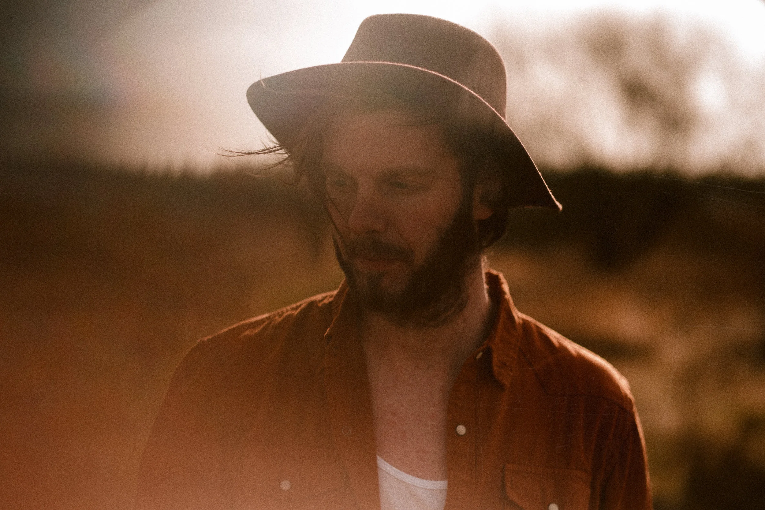 A man with dark hair, beard, glasses, wearing a wide-brimmed hat and a reddish-orange shirt, looking downward in a sunny outdoor setting with blurred background.
