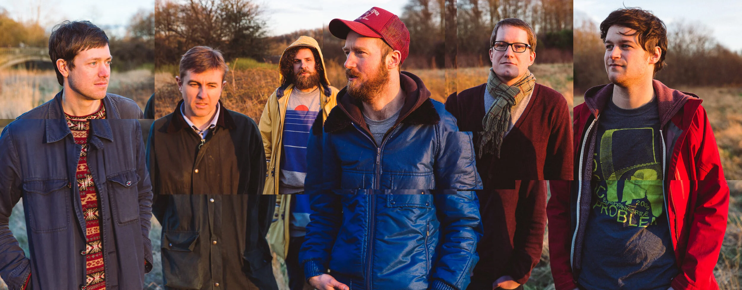 Group of seven men standing outdoors in a field during sunset, wearing casual jackets and sweaters, some with scarves and hats, with autumn trees in the background.