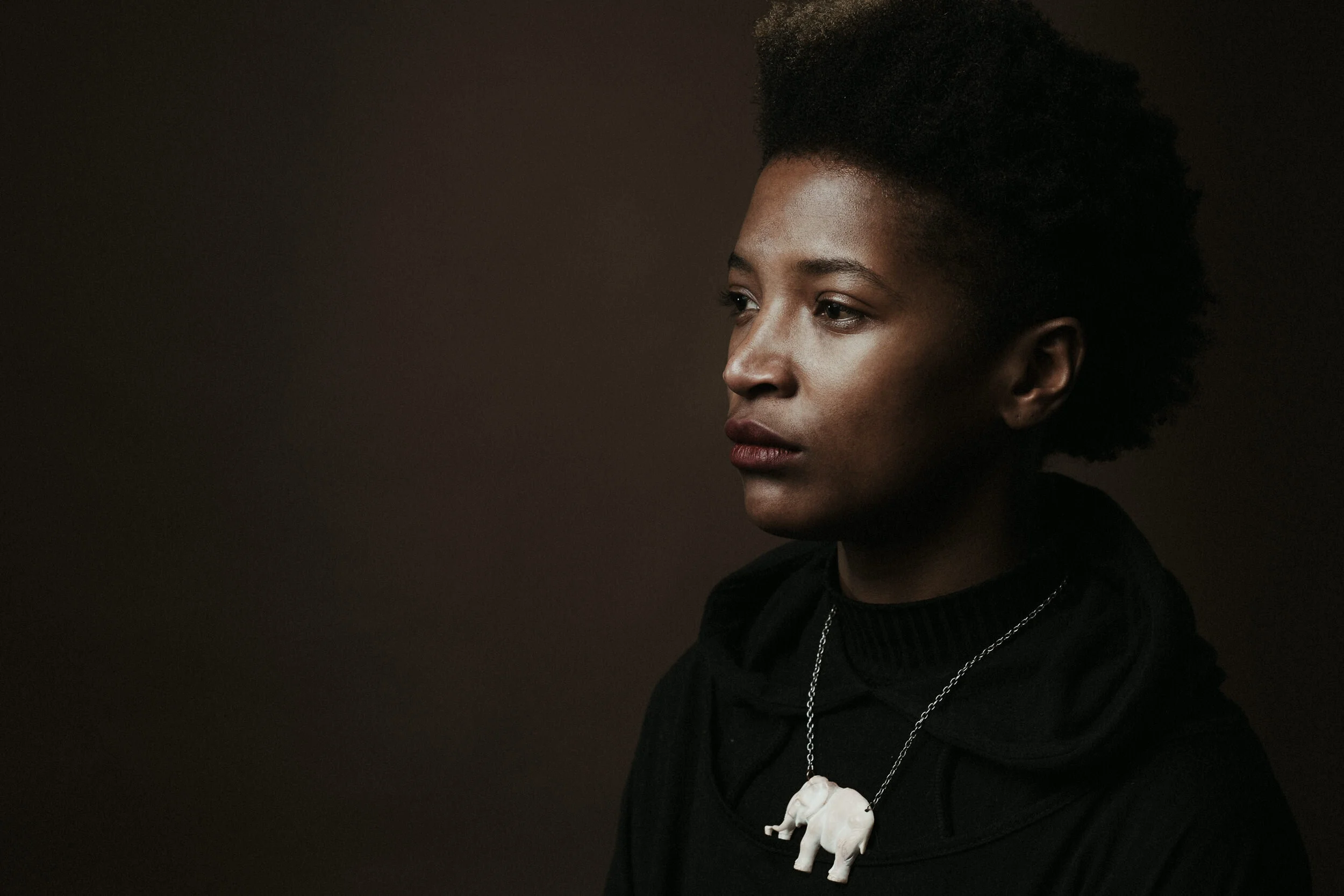 Portrait of a woman with short, natural hair against a dark background, wearing a black top and a white skull necklace.