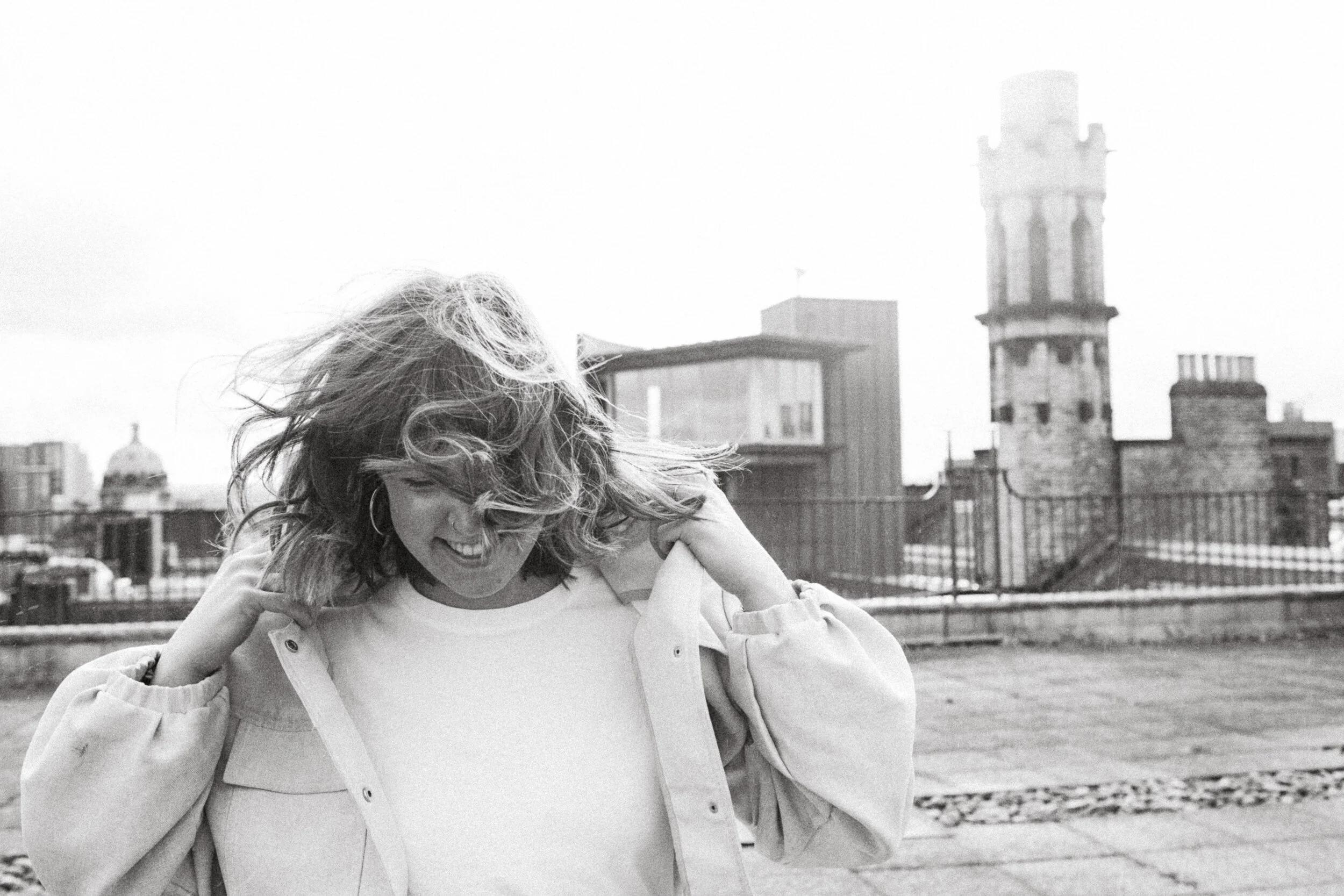 A woman with curly hair smiling and adjusting her jacket on a rooftop with city buildings and a tower in the background.