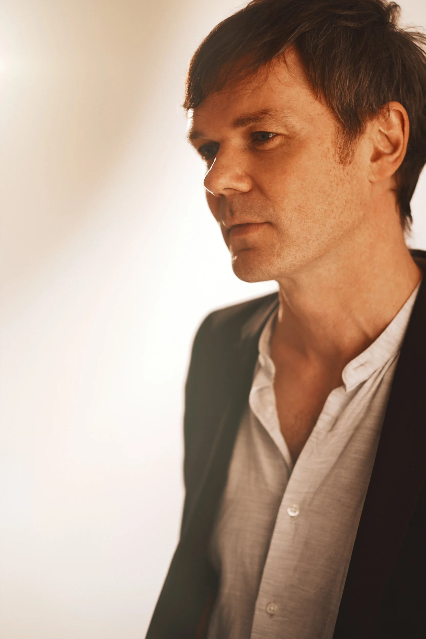 A close-up profile of a man with dark hair, wearing a gray blazer and white shirt, looking downward against a plain background.