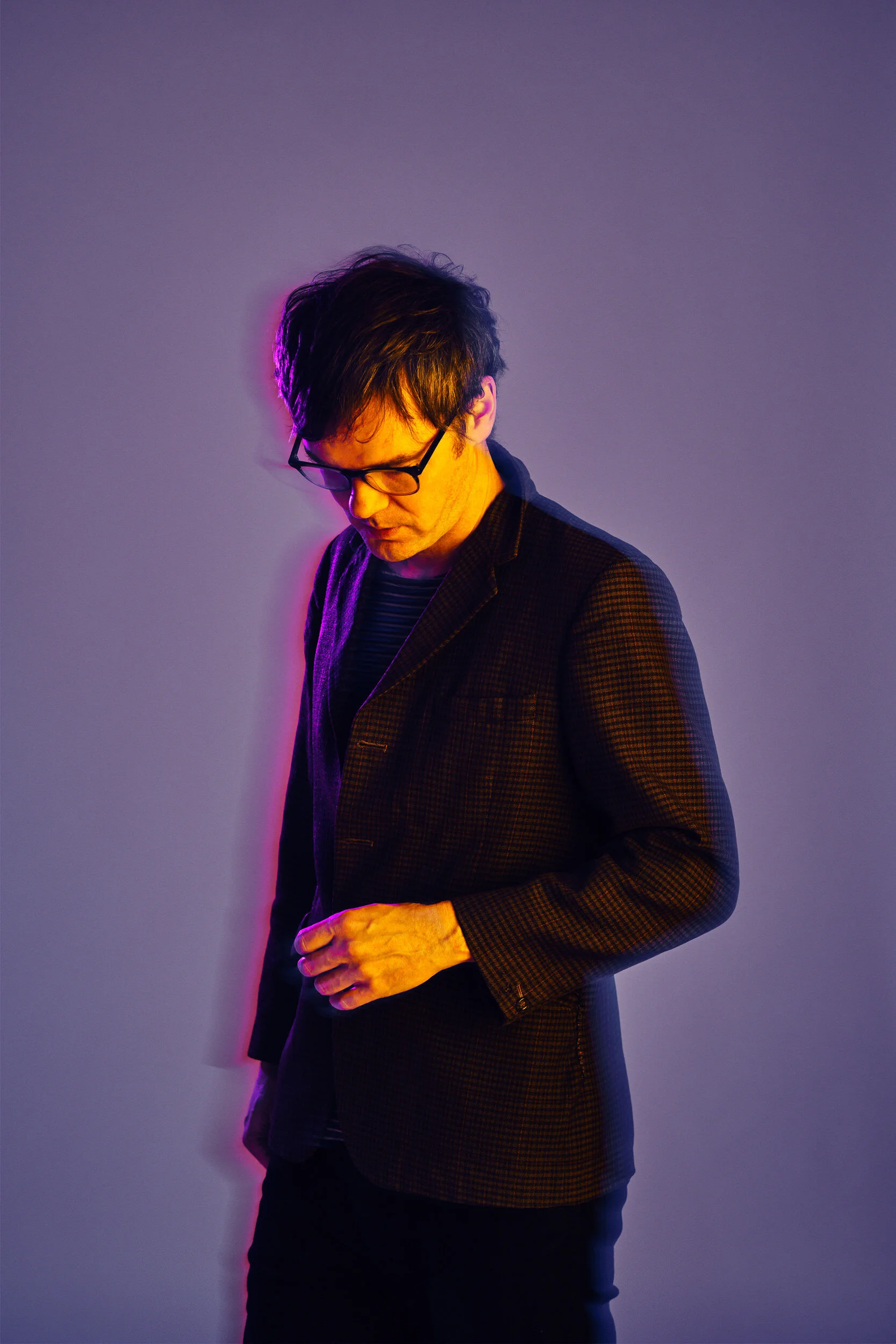 A man with dark hair, glasses, and wearing a checkered blazer over a striped shirt, stands against a plain wall with purple lighting. He looks down, with one hand near his waist.