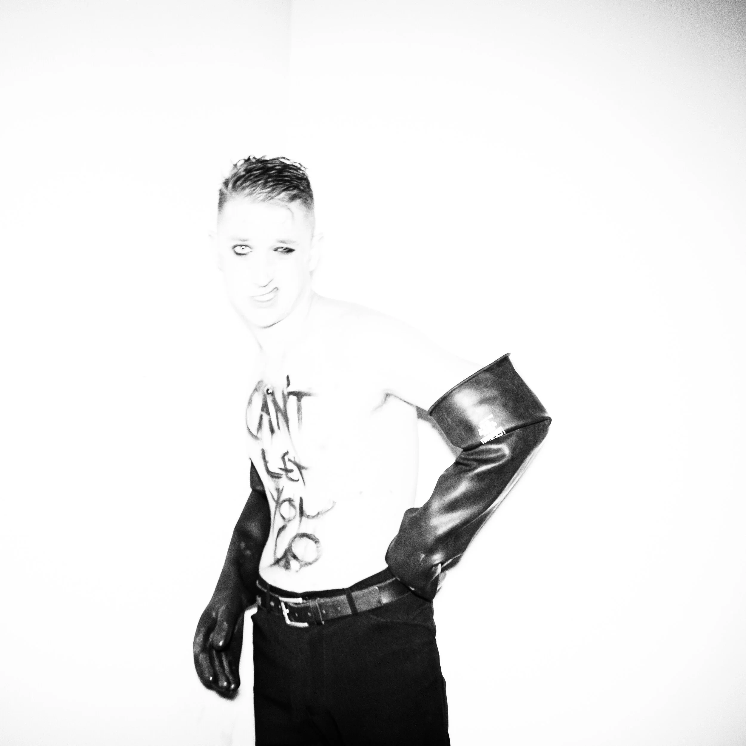 Black and white photo of a young man with short hair, wearing a T-shirt, leather jacket, and jeans, holding a cup, standing against a plain background.