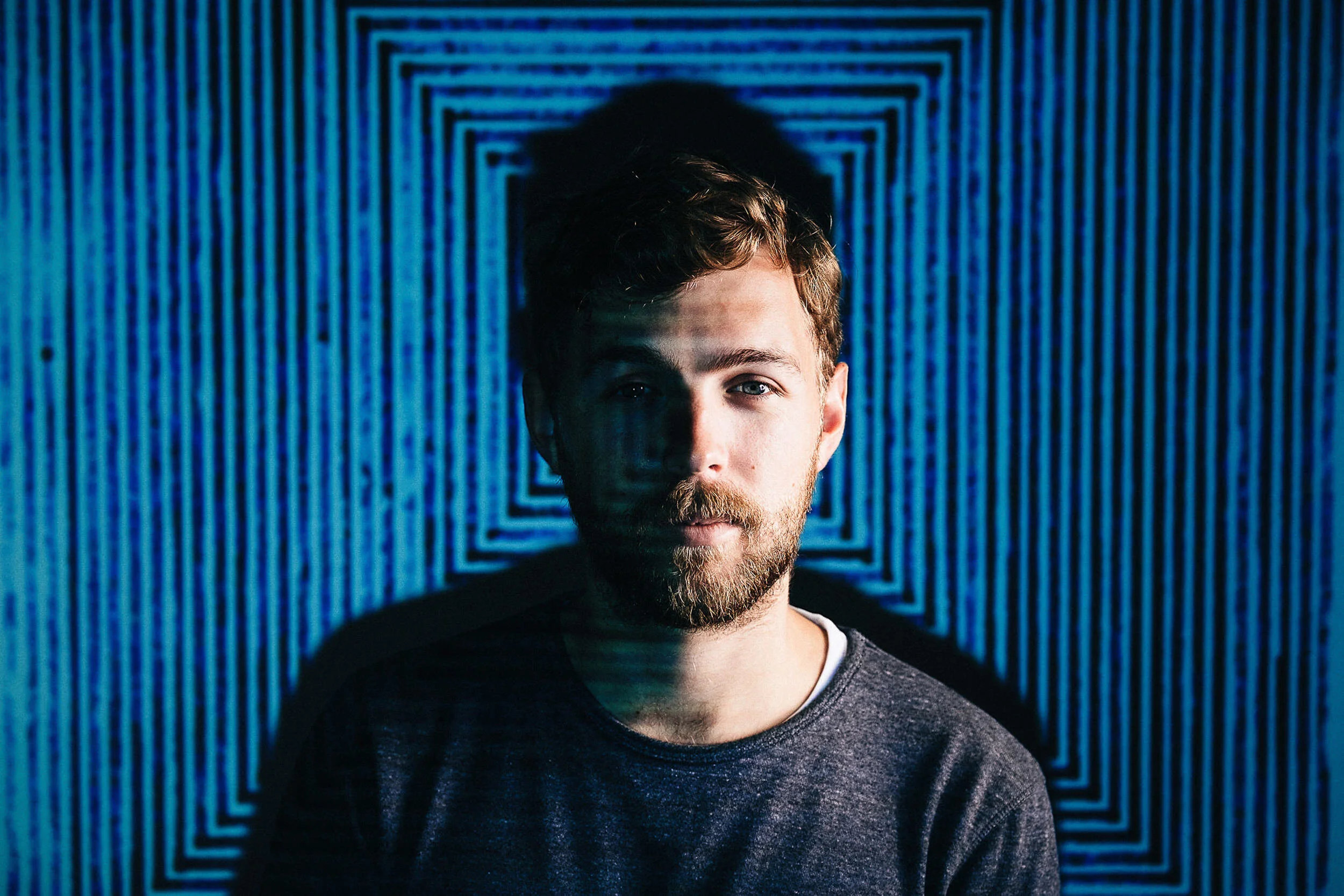 A young man with light brown hair and a beard, wearing a gray shirt, stands in front of a geometric blue light pattern inside a tunnel or corridor.