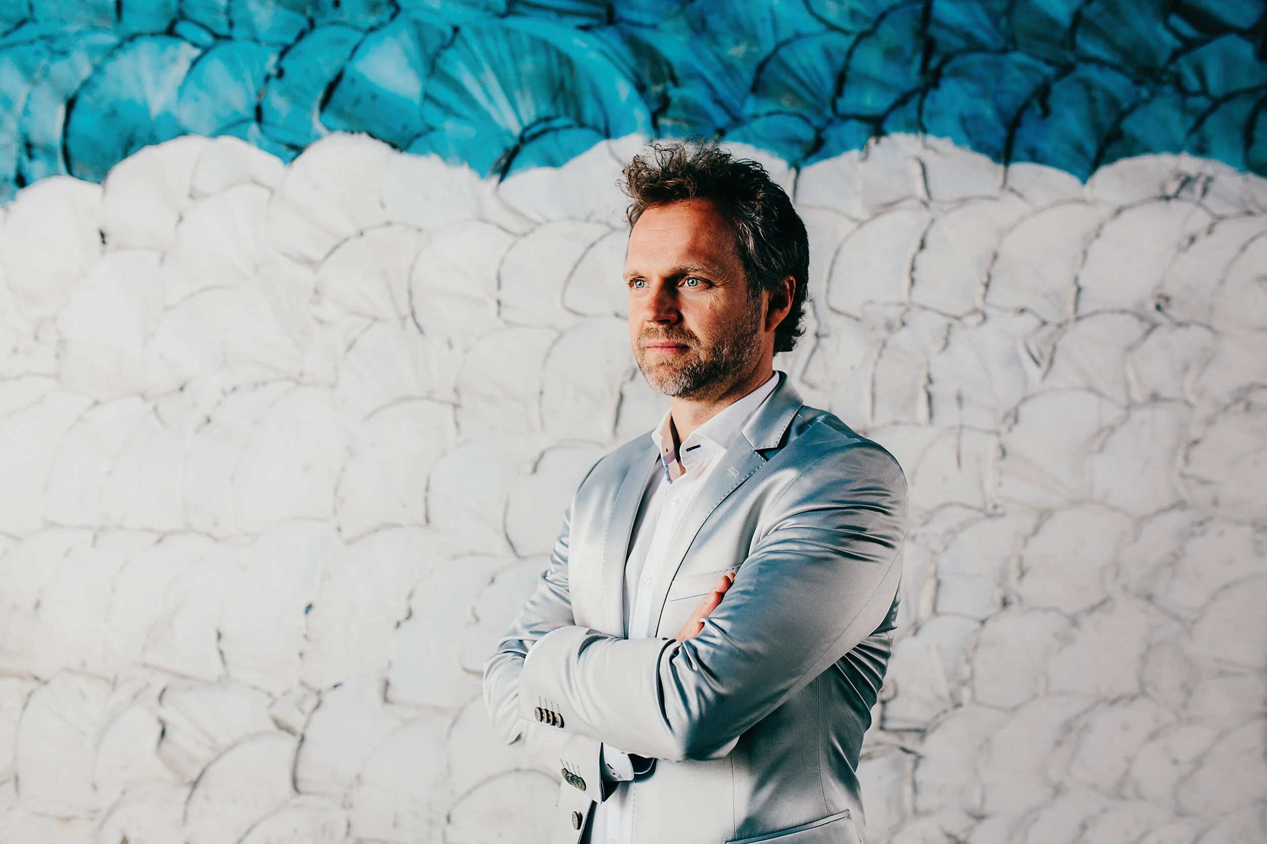 A man with blue eyes and dark curly hair, wearing a light grey suit and white shirt, stands with his arms crossed in front of a white stone wall with blue abstract art painted on it.