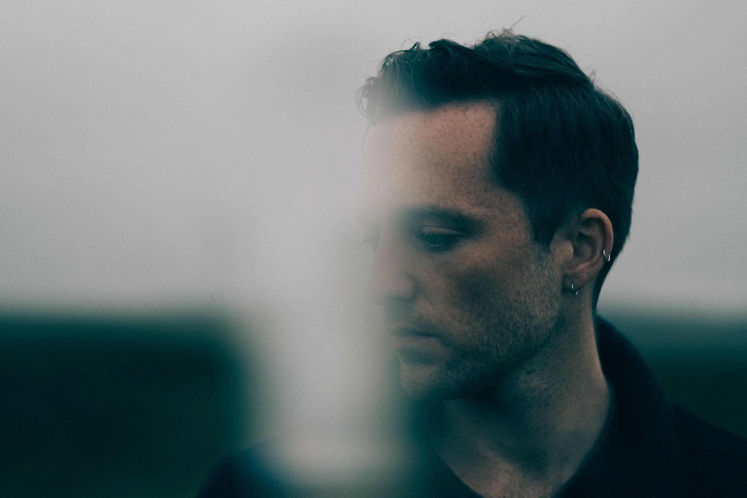Close-up of a young man's face partially visible with short dark hair, wearing earrings, against a blurred background.