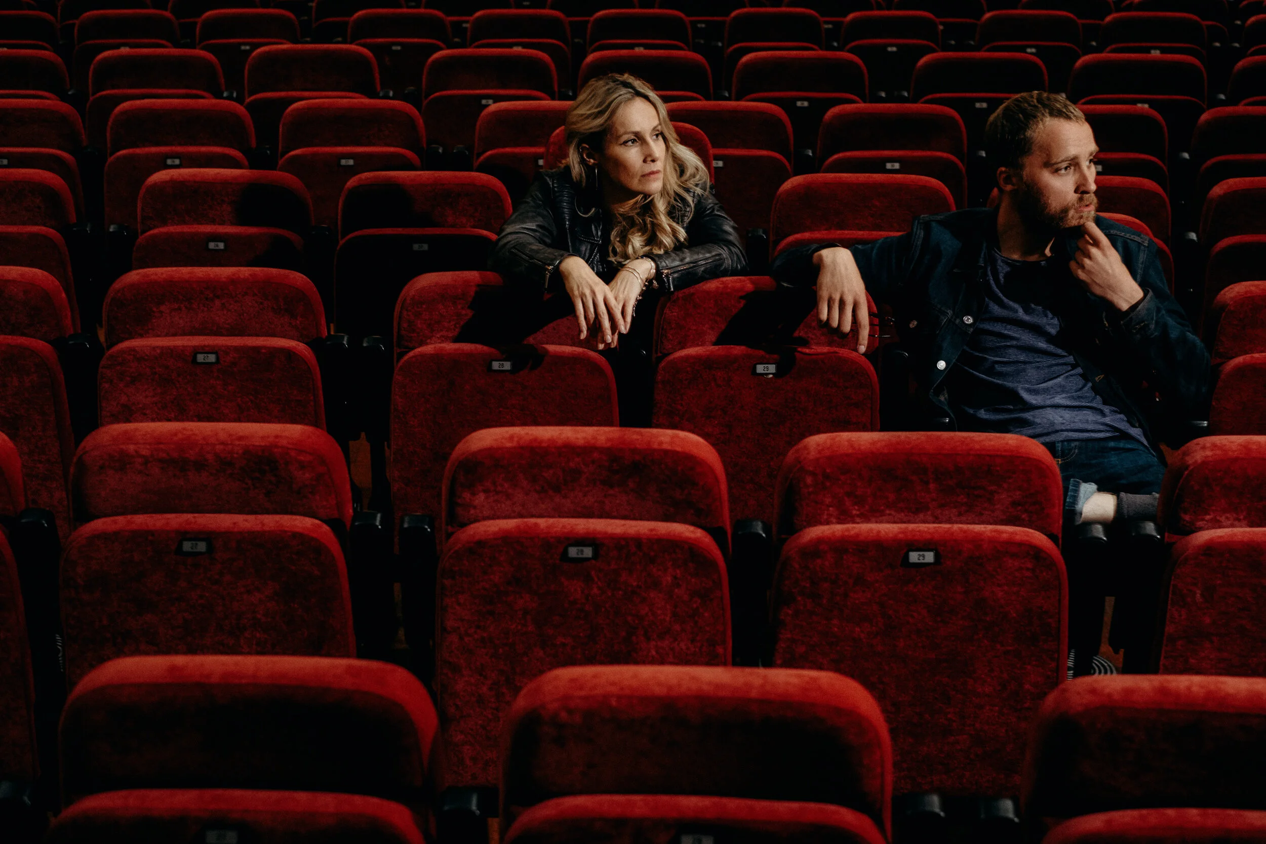 A woman and a man sitting in an empty theater with red velvet seats. The woman is leaning on her arm looking to her right, while the man is sitting with his hand on his chin, also looking to the right.